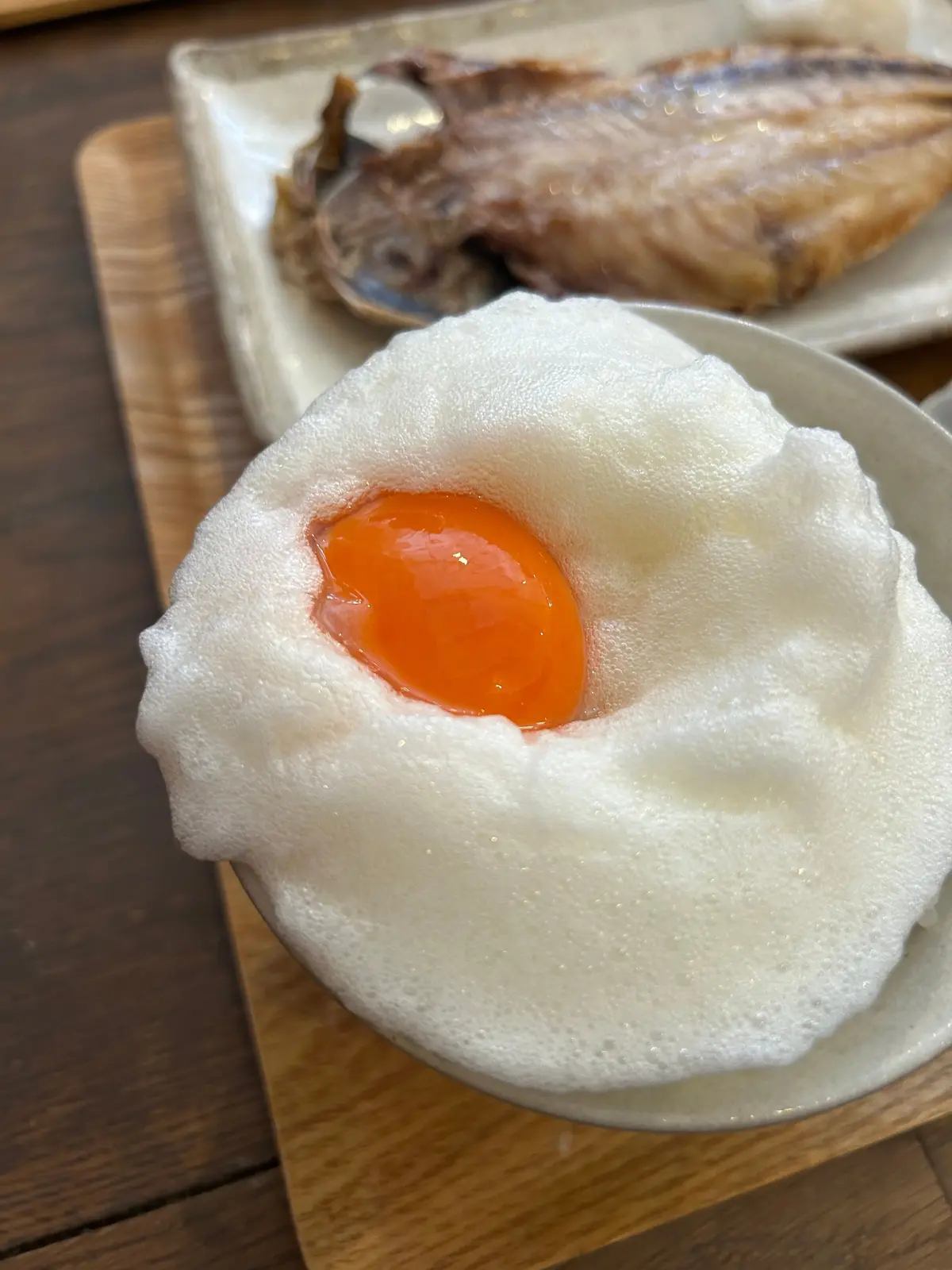 白い泡に包まれた半熟卵。黄身は鮮やかなオレンジ色で、中央に位置している。背景には、白い皿に盛り付けられた魚料理が見える。木製のトレイに置かれている。