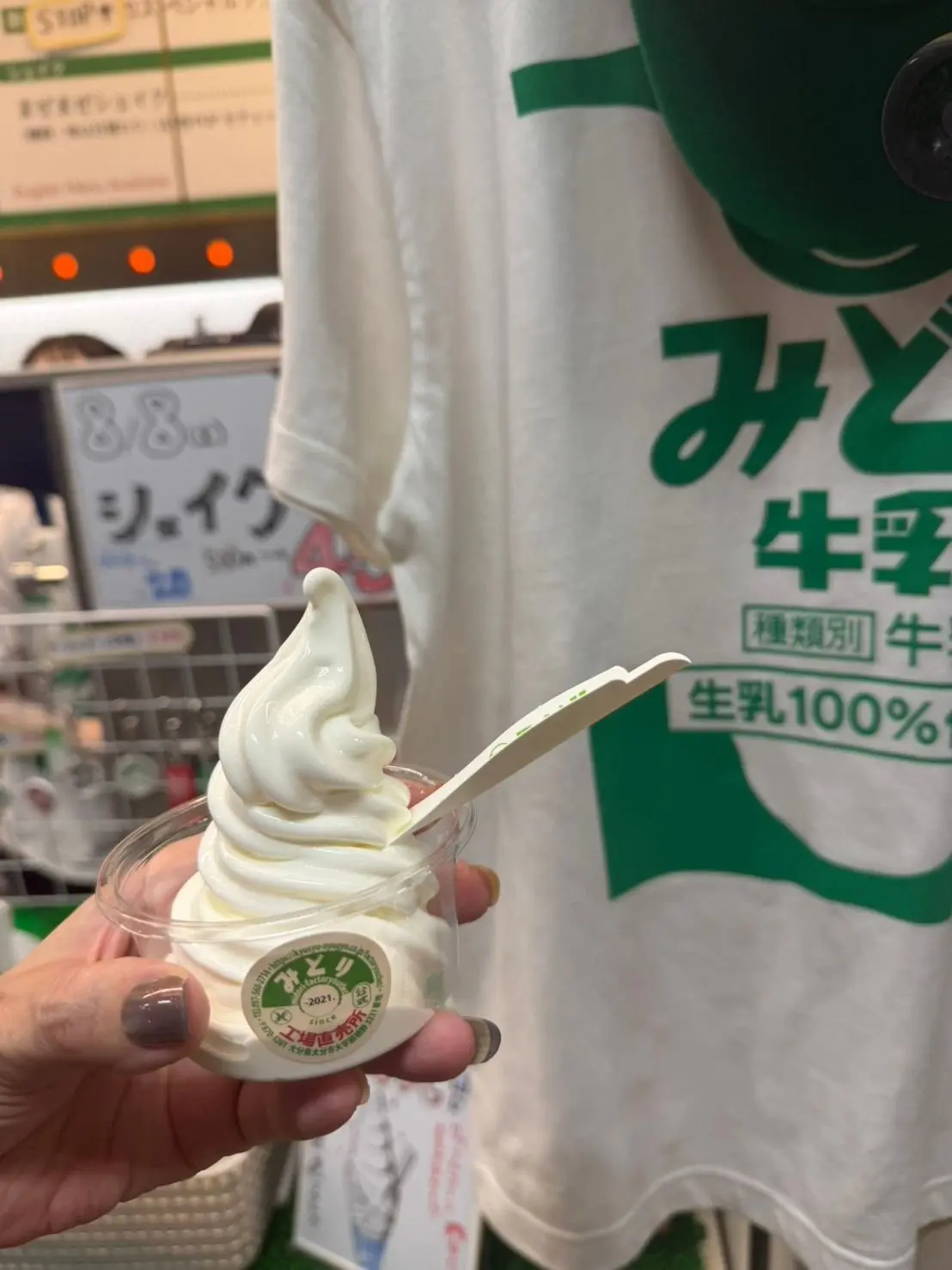透明なカップに入ったソフトクリーム。白いクリームが渦巻き状に盛り付けられ、緑色のスプーンが挿さっている。背景には「みやぞの」のロゴと、ソフトクリームの看板が見える。