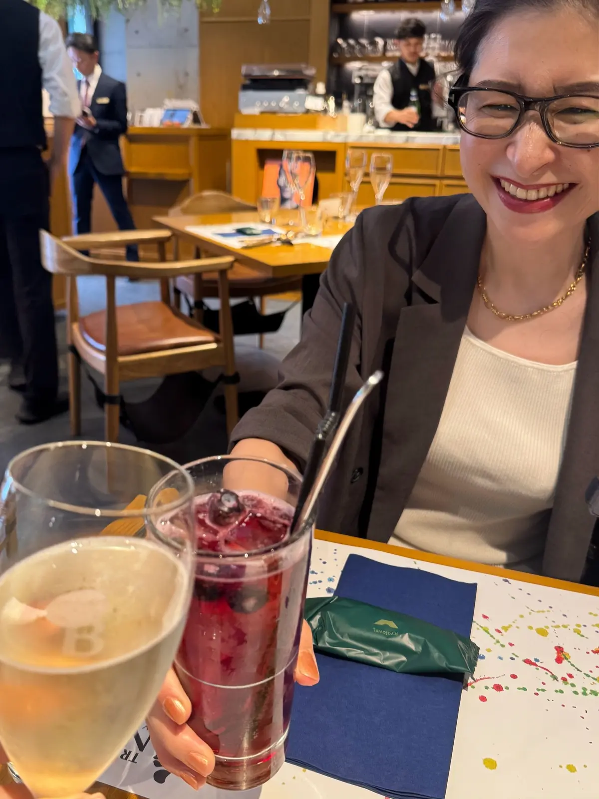 表参道レストラン、イタリアンレストラン庭、メガネをかけた女性が、赤いカクテルと白い飲み物を含むテーブルに座り、笑顔でカメラを見ている。背景にはカウンターと他の客人が見える。