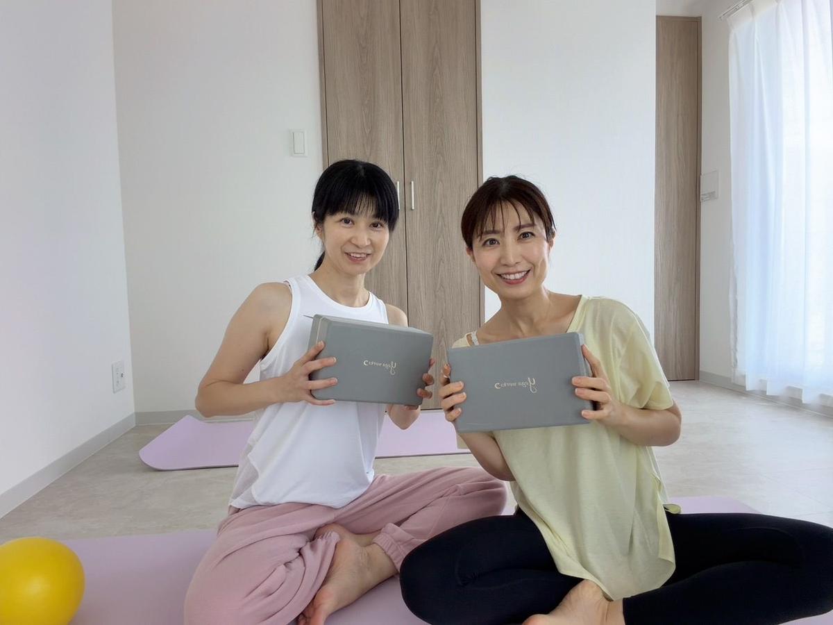 ヨガマットの上に座った2人の女性が、グレーの箱型の製品を笑顔で持っている。背景には白いカーテンと窓が見える。