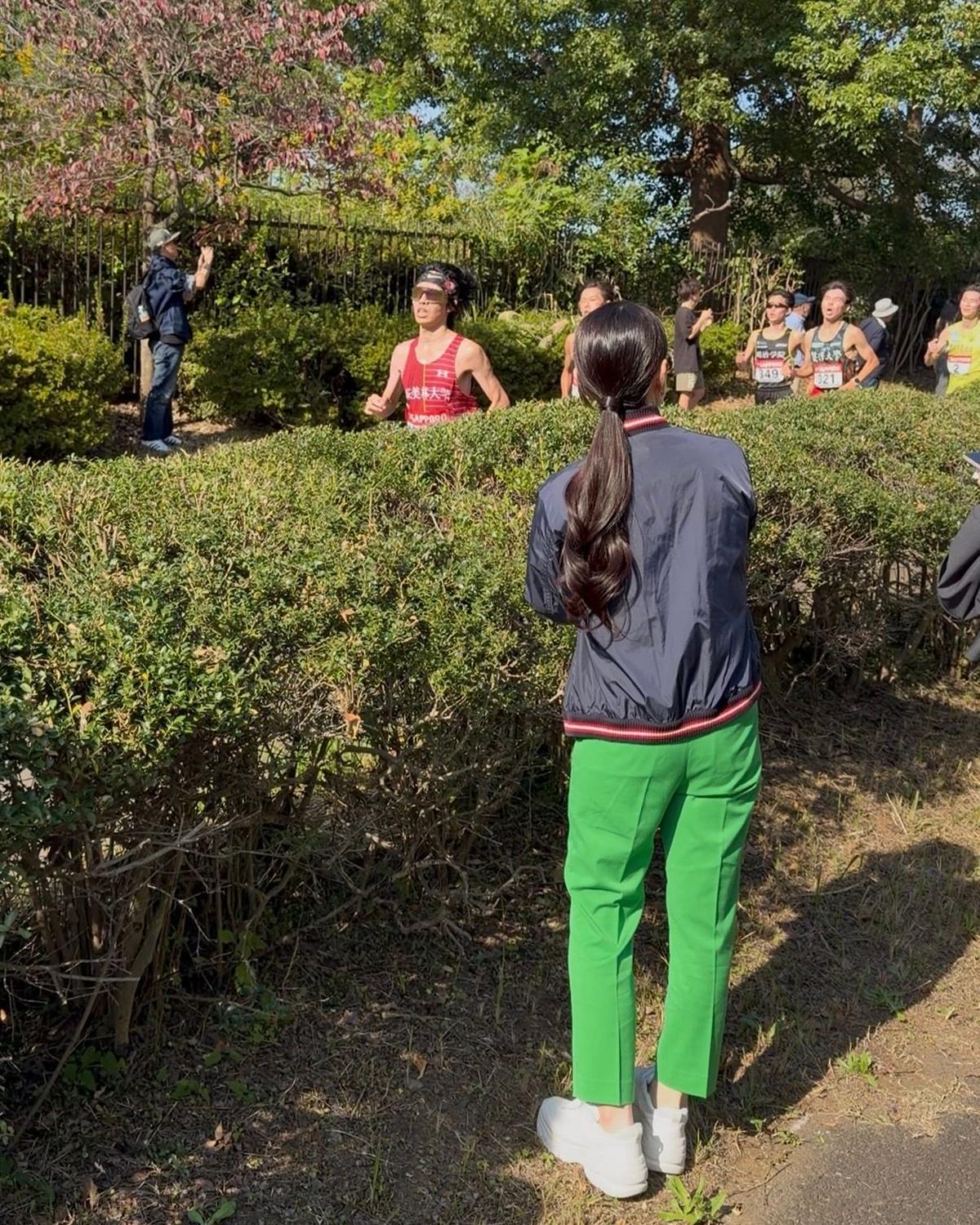 屋外のレース中、赤いゼッケンをつけた男性ランナーが先頭を走る。背景には観客や他のランナーが見える。女性が緑色のズボンと紺色のジャケットを着て、その様子を見ている。