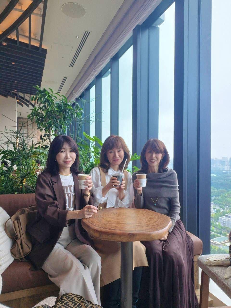 3人の女性が、窓際に置かれた木製の丸テーブルを囲んで座り、それぞれコーヒーカップを持っている。女性たちは笑顔で、カジュアルな服装をしている。背景には高層ビルの窓と緑が見える。