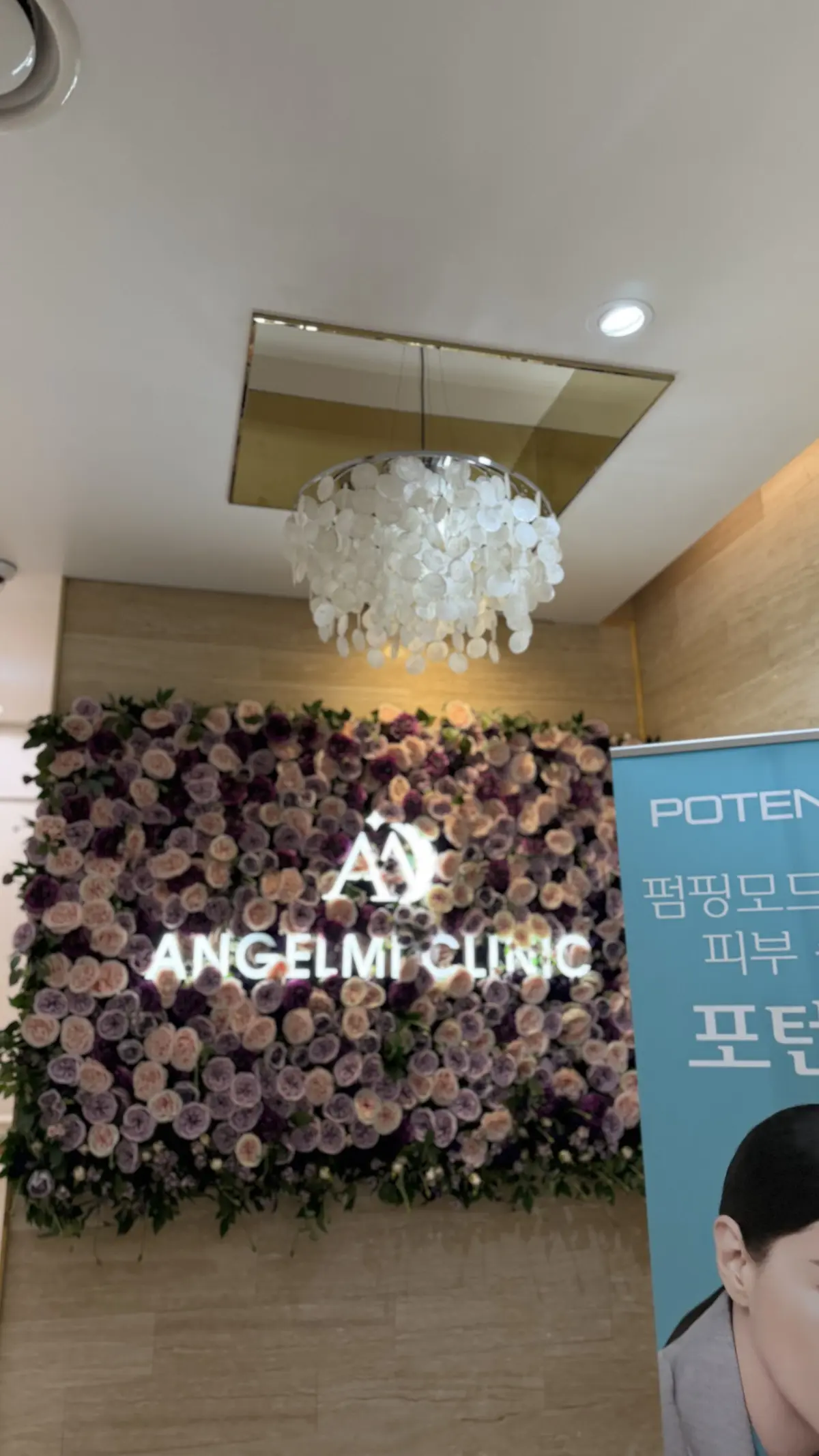 アンジェルメクリニックのロゴマークが中央に配置された、紫色の花でできた壁飾り。上部には白いシャンデリアが吊り下げられている。右側には青色のポスターが貼られている。