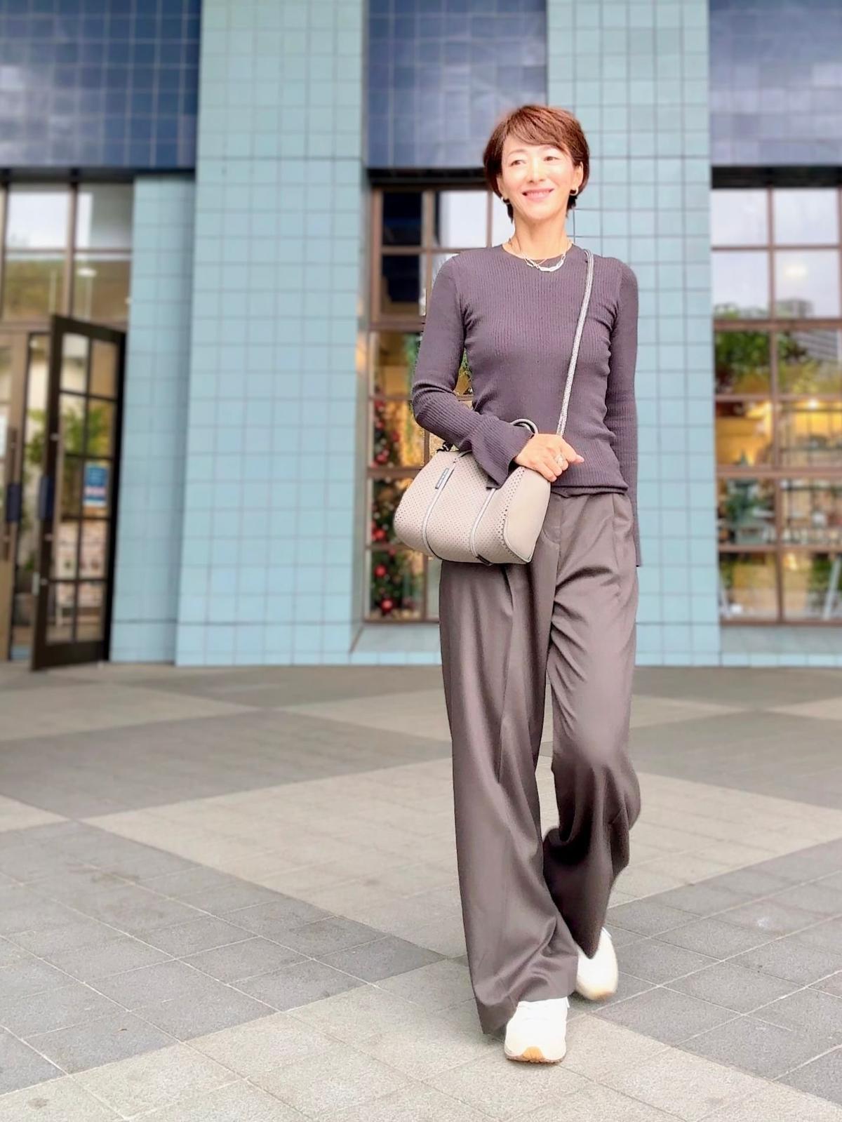グレーのトップスとパンツを着用した女性が、灰色のコンクリートの歩道で歩いている。右手に白のバッグを持ち、笑顔を浮かべている。背景には淡いブルーの建物がある。