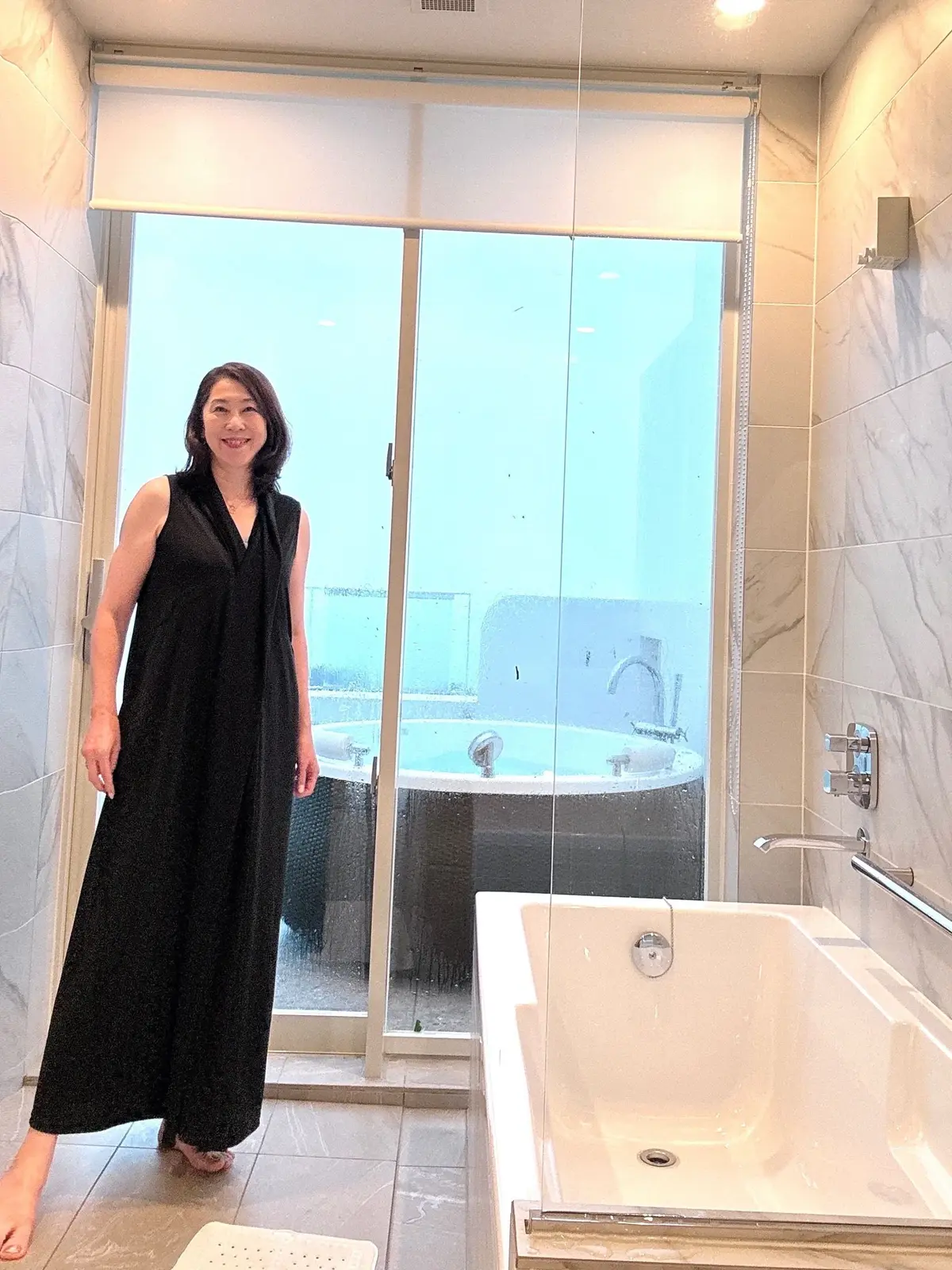 黒いワンピースを着た女性が、白いタイル張りのバスルームに立っている。背後にはガラスの引き戸があり、外の景色が見える。手前に白いバスタブ、右にはシャワー設備がある。