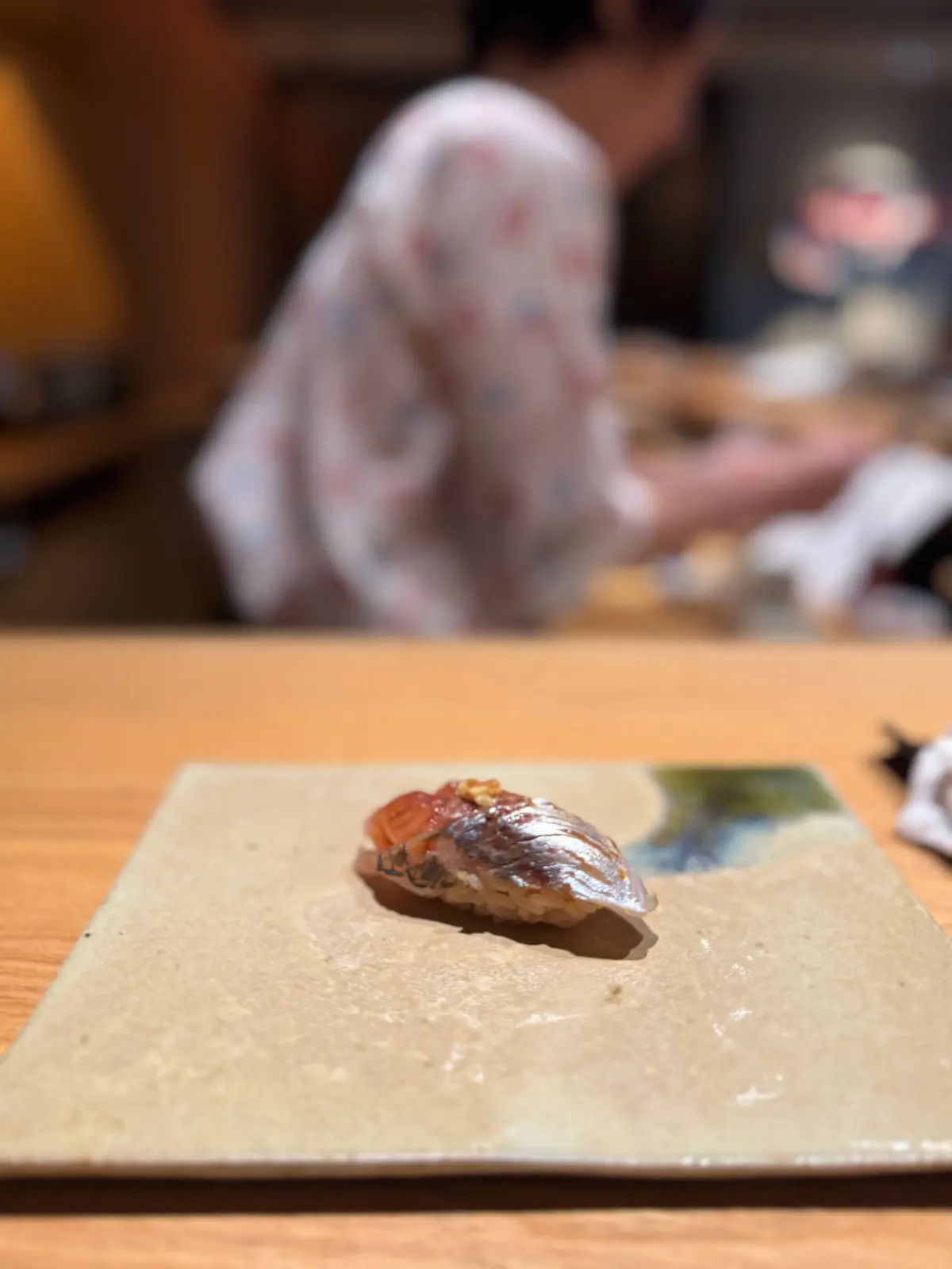 寿司一貫。赤身の魚の上にオレンジ色の層があり、白米の上に置かれている。木製の皿の上で、背景には寿司職人がいる。