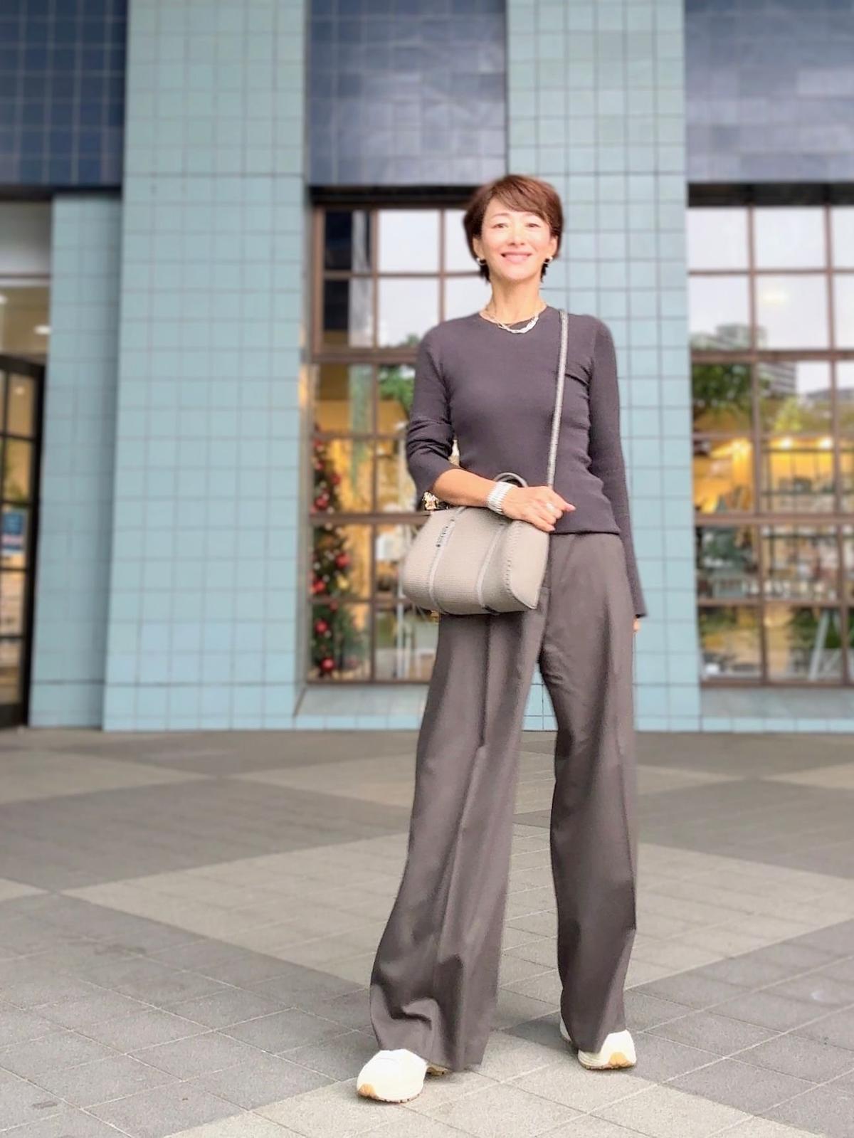 グレーの長袖ニットとワイドパンツを着用した女性が、建物の前でポーズをとっている。右手にグレーのショルダーバッグを持ち、白いスニーカーを履いている。背景は青色のタイル張りの壁と窓。