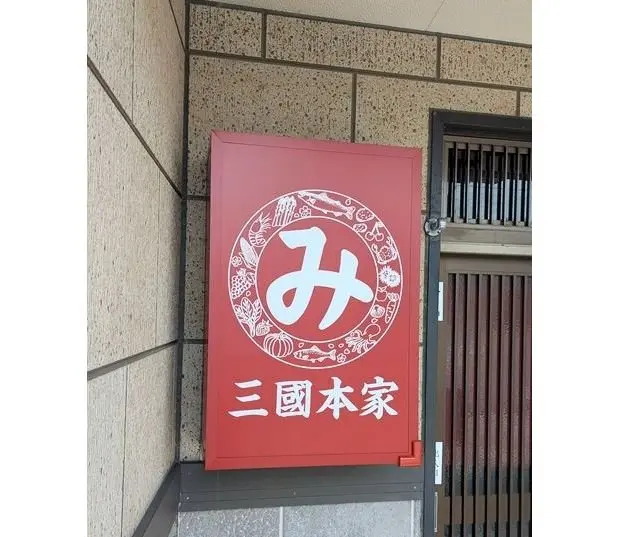 赤色の長方形の看板に、中央に白文字の「み」の文字と、その周りに様々な食材のイラストが描かれている。下部には白文字で「三国本家」とある。