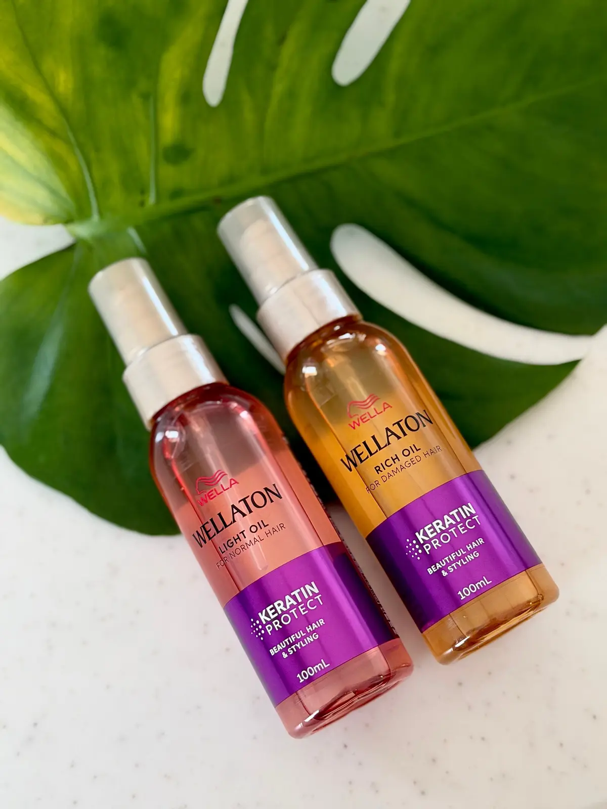 白いテーブルに置かれた、ウェラ（Wella）のヘアオイル2本。左は「ライトオイル」、右は「リッチオイル」。それぞれ紫色のラベルに「Keratin Protect」のロゴと100mlの容量表記がある。背景には緑の葉。