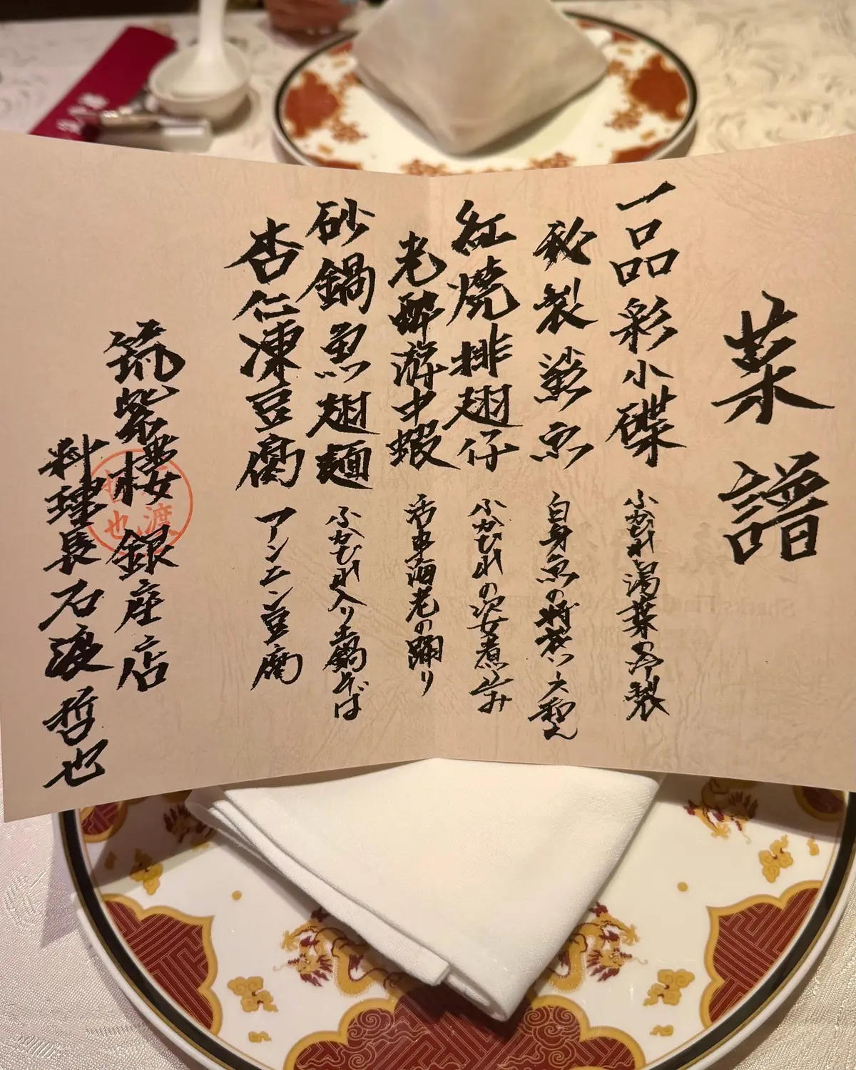 手書きのメニューが茶色い紙に記載され、伝統的な中国料理の品名が並んでいる。背景には白い布ナプキンと金色の装飾が施された陶器の皿が置かれている。