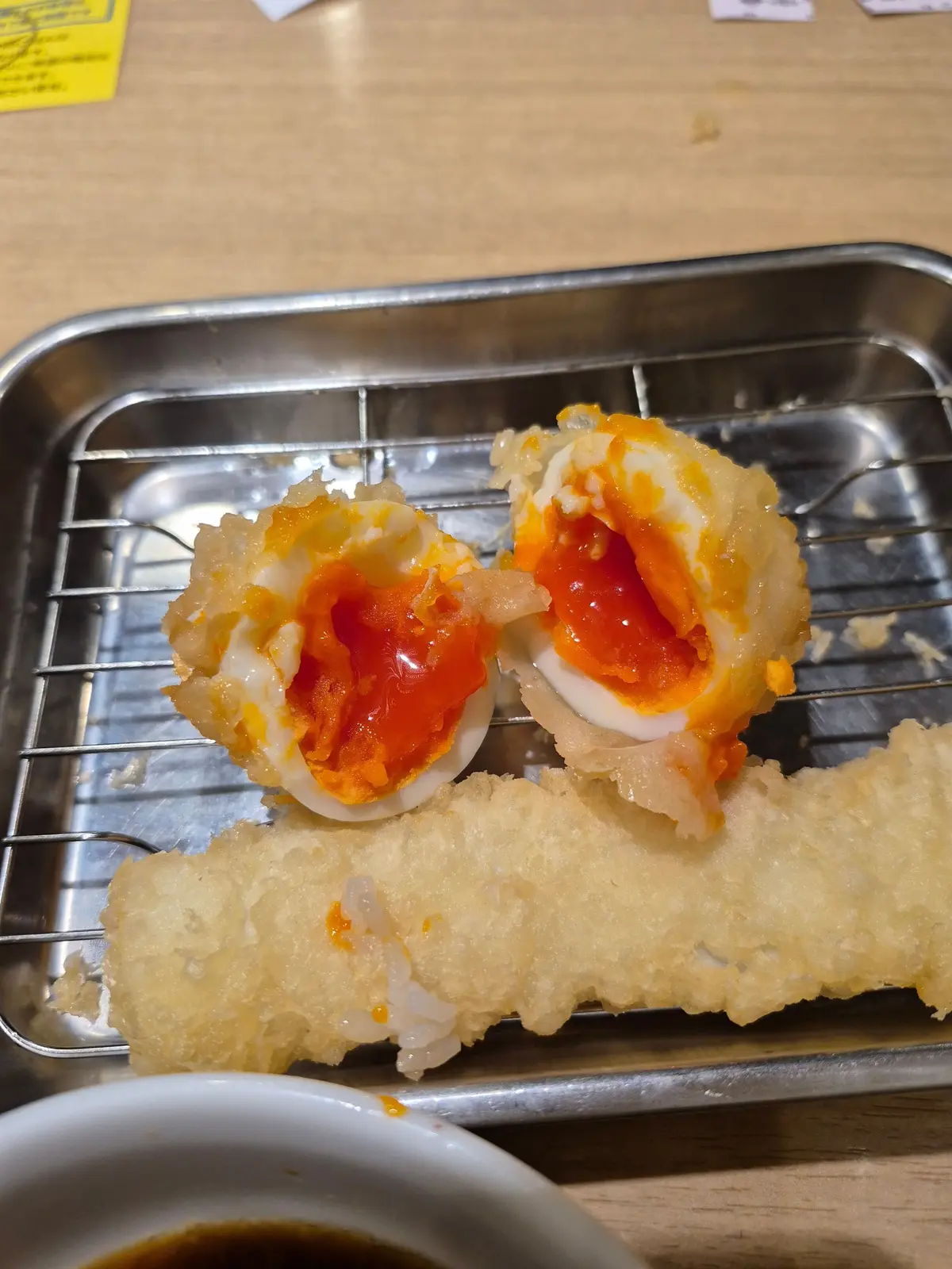 金属製のトレイに、天ぷら衣で包まれた半熟卵が3つ。2つは縦に半分にカットされ、中から赤い卵黄が見える。手前には、衣の一部が割れた天ぷらが置かれている。