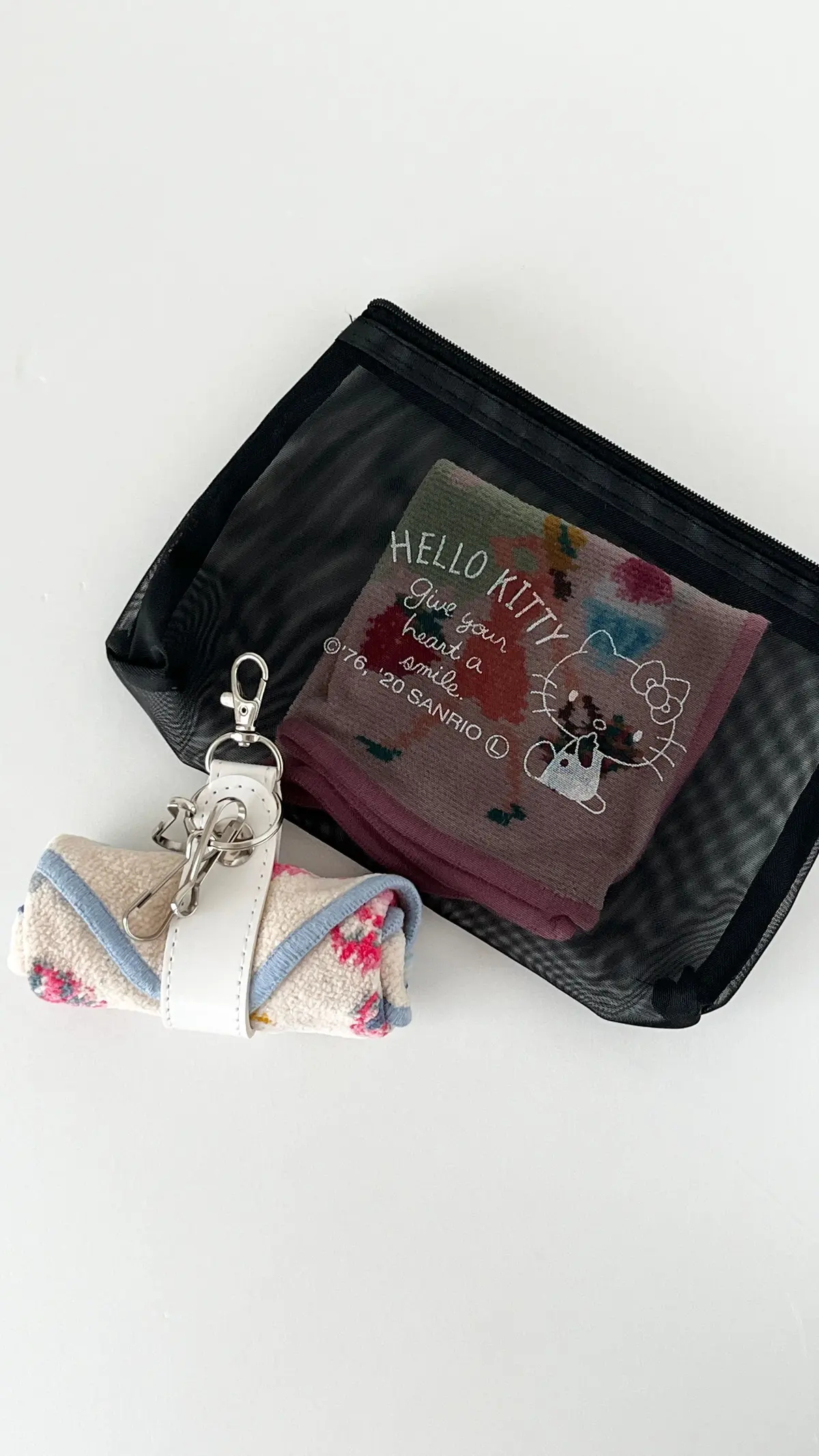 黒いメッシュ素材のポーチに入った、Hello Kittyのハンカチが映る写真。ハンカチにはHello Kittyのイラストと「Give your heart a smile.」の文字がプリントされている。ポーチにはキーホルダーも付いている。