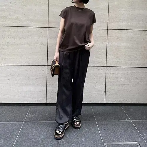 茶色のTシャツに黒いワイドパンツ、黒いサンダルを合わせた女性。右手に茶色の小さなバッグを持ち、正面を向いている全身写真。背景はグレーのタイル壁。