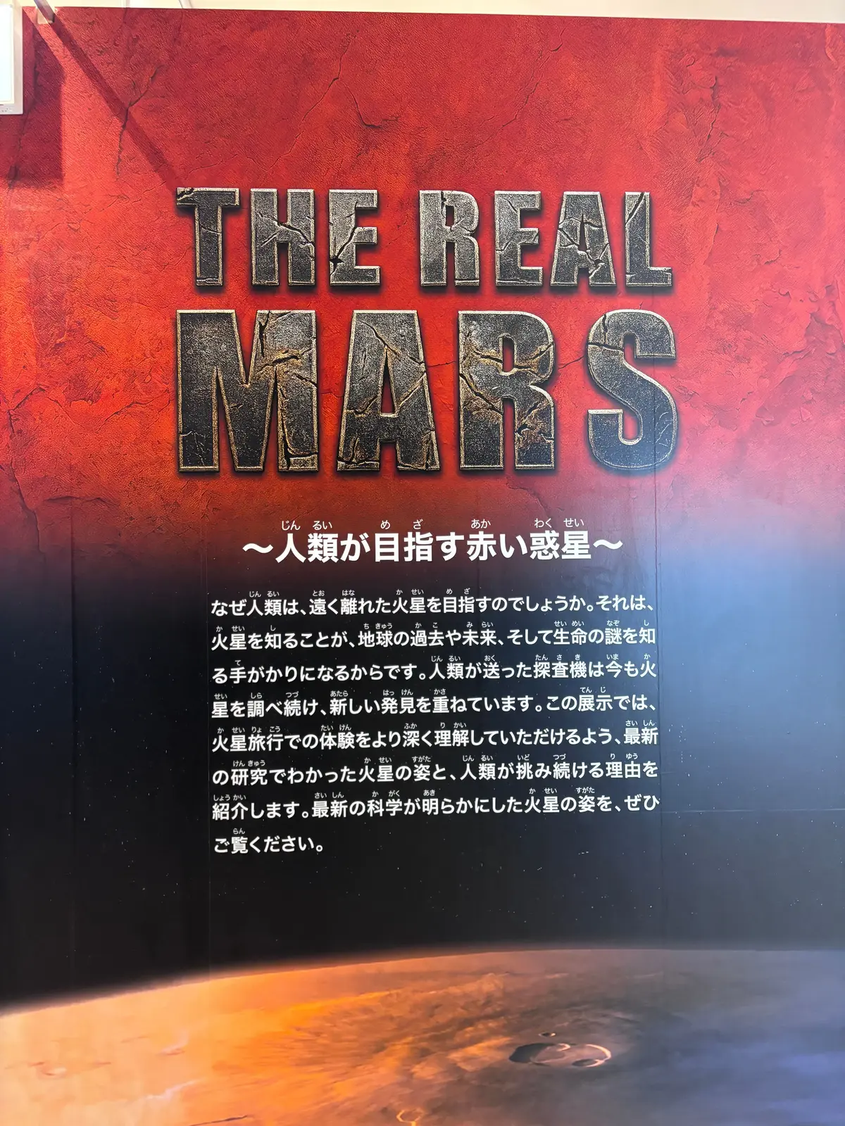 赤い砂漠の風景を背景に、黒い文字で「THE REAL MARS」と大きく書かれたポスター。下部には日本語で「人類が目指す赤い惑星」などのテキスト。画面下部には、火星探査機のイメージ。