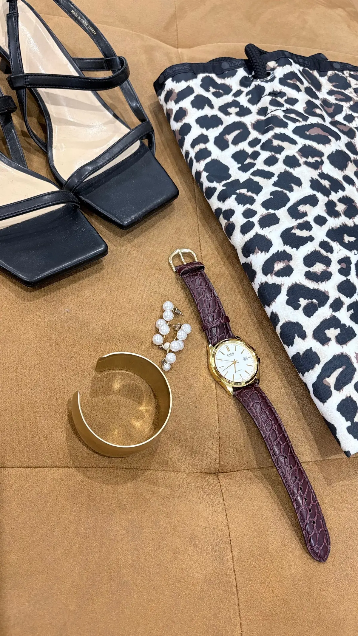 leopard柄のクラッチバッグ、ゴールドの腕時計、真珠のイヤリング、ゴールドの腕輪、ネイビーのサンダルが茶色の背景に並んでいる