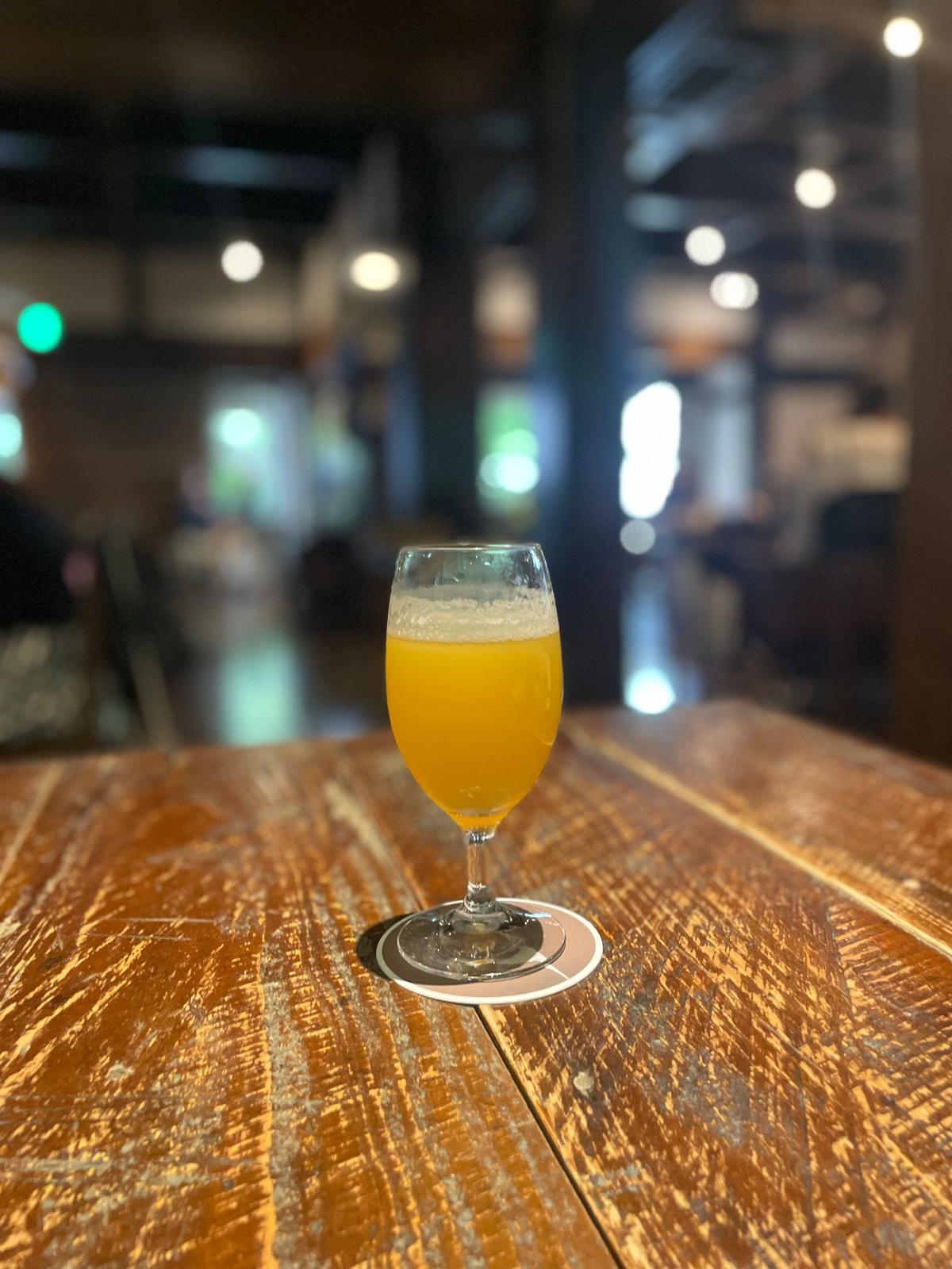 木製のテーブルに置かれた、オレンジ色のビールが入ったグラス。表面に白い泡が乗っており、背景にはぼやけた照明と人々が見える。