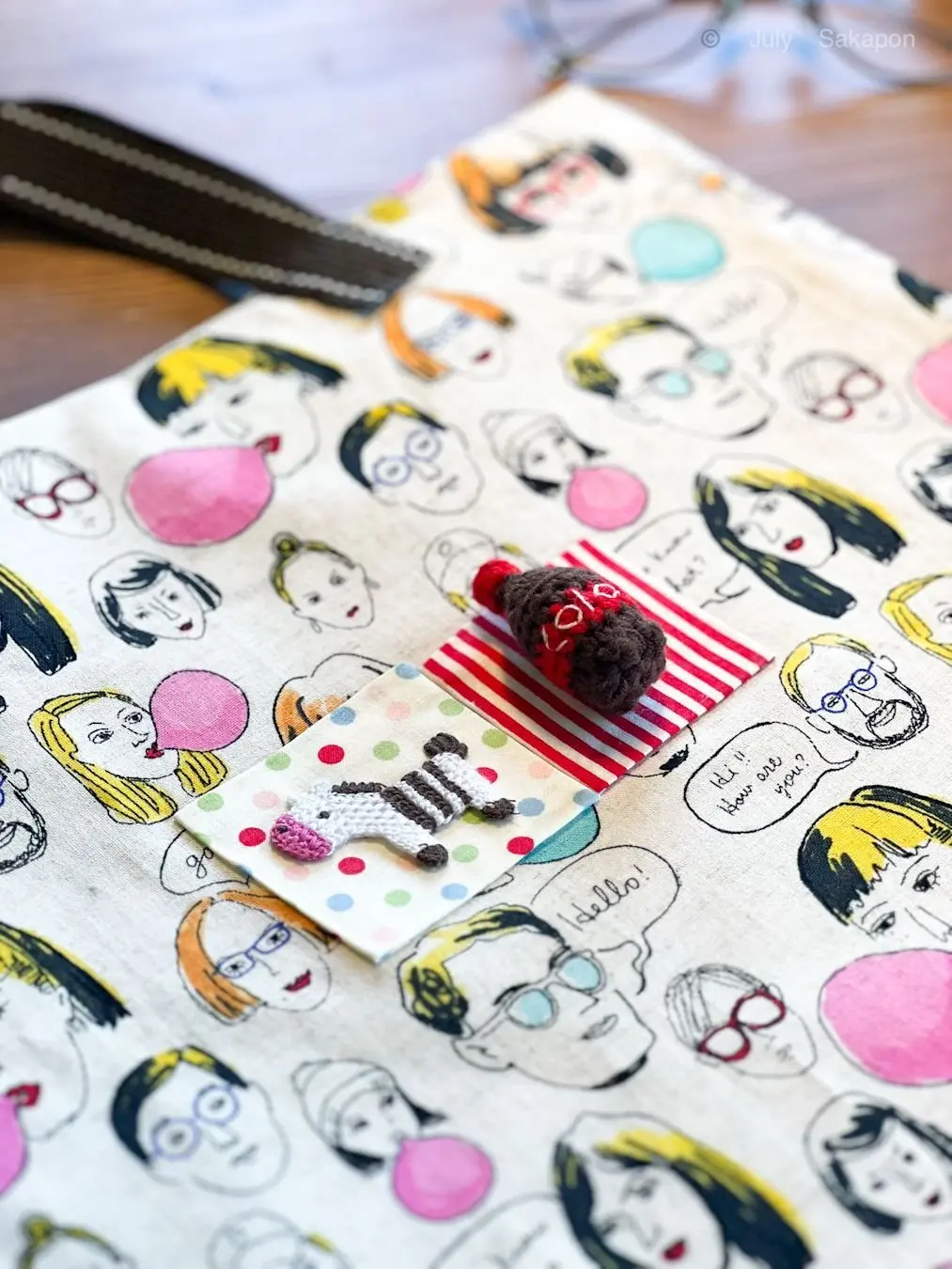 手作りの小さな人形が、キャラクターが描かれた布製のノートの上に置かれている。人形は赤と白の縞模様で、顔には「LOVE」と書かれている。ノートには様々なキャラクターのイラストと会話風のテキストが印刷されている。
