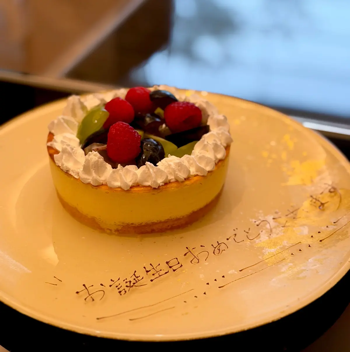 黄色の皿に置かれた、丸いレモンタルト。表面にはホイップクリームと、キウイ、ラズベリー、ブルーベリーなどのベリー類が飾られている。皿には日本語でメッセージが書かれている。