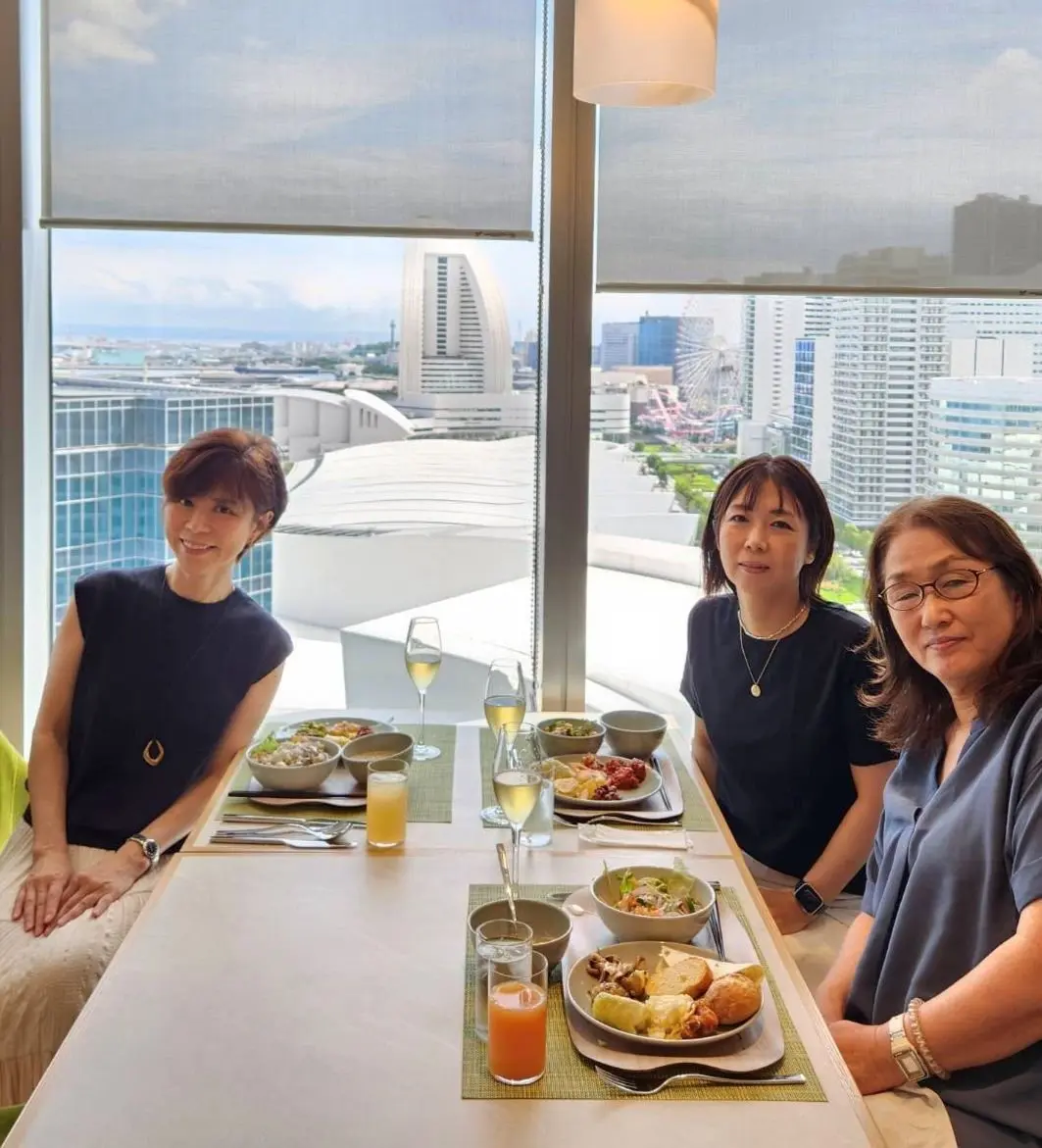高層階のレストランで、3人の女性がテーブルを囲んで食事を楽しんでいる様子。窓からは都市の景色が見える。テーブルにはサラダや料理、飲み物が並び、明るい雰囲気。