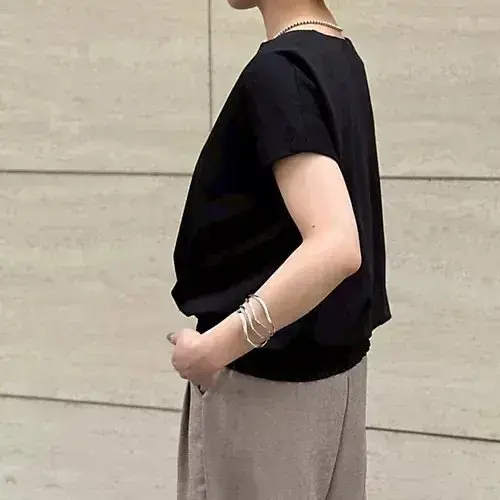 黒の半袖シャツとグレーのスカートを着た女性が、左手でスカートを少し持ち上げ、右手には複数の銀のブレスレットが光る。背景はベージュのコンクリート壁。