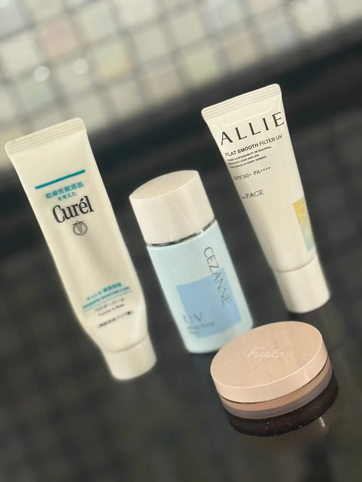 3つのスキンケア製品とパウダー容器が並んでいる。左から「Curel」のクリームチューブ、中央に「CEZANE」の青いボトル、右に「ALLIE」のフェイスクリームチューブと、茶色の円形パウダーケース。