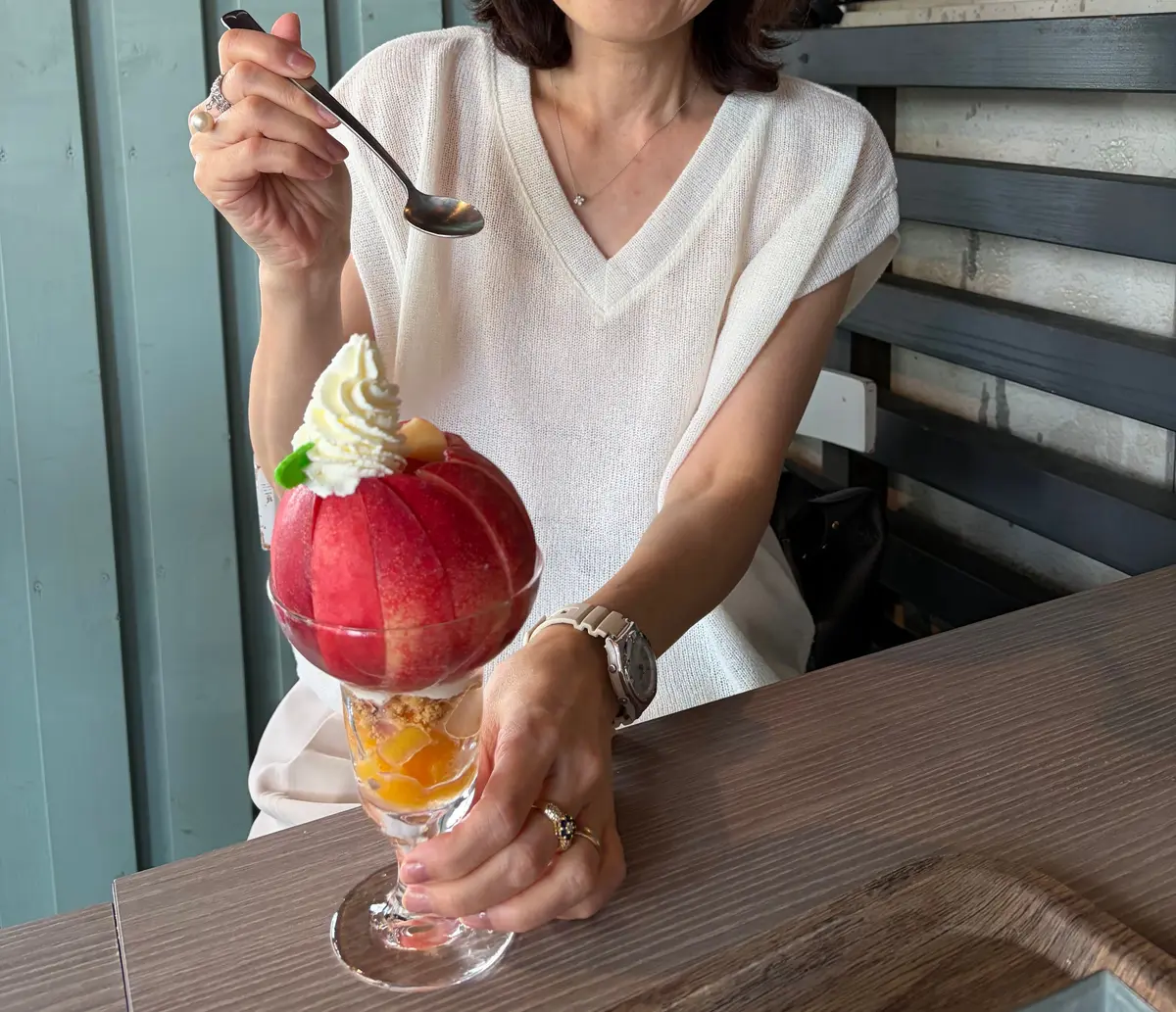 白いVネックのトップスを着た女性が、ガラスの器に入ったフルーツゼリーをスプーンで混ぜている。背景には木製の壁と窓があり、植物が見える。