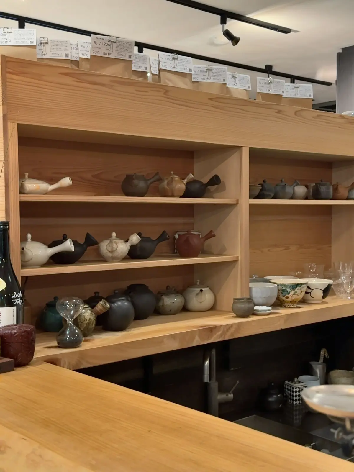 木製の棚に、さまざまな形の茶器が並んでいる。茶器は白、茶色、黒などの色で、大小さまざまなサイズがある。棚の上には価格表示のタグが吊るされている。