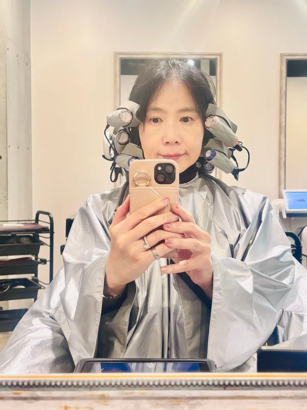 美容室でヘアドライヤーの温風機を装着した女性が、スマートフォンで自撮りをしている。銀色の保護シートを着用し、前髪には複数の温風機が配置されている。