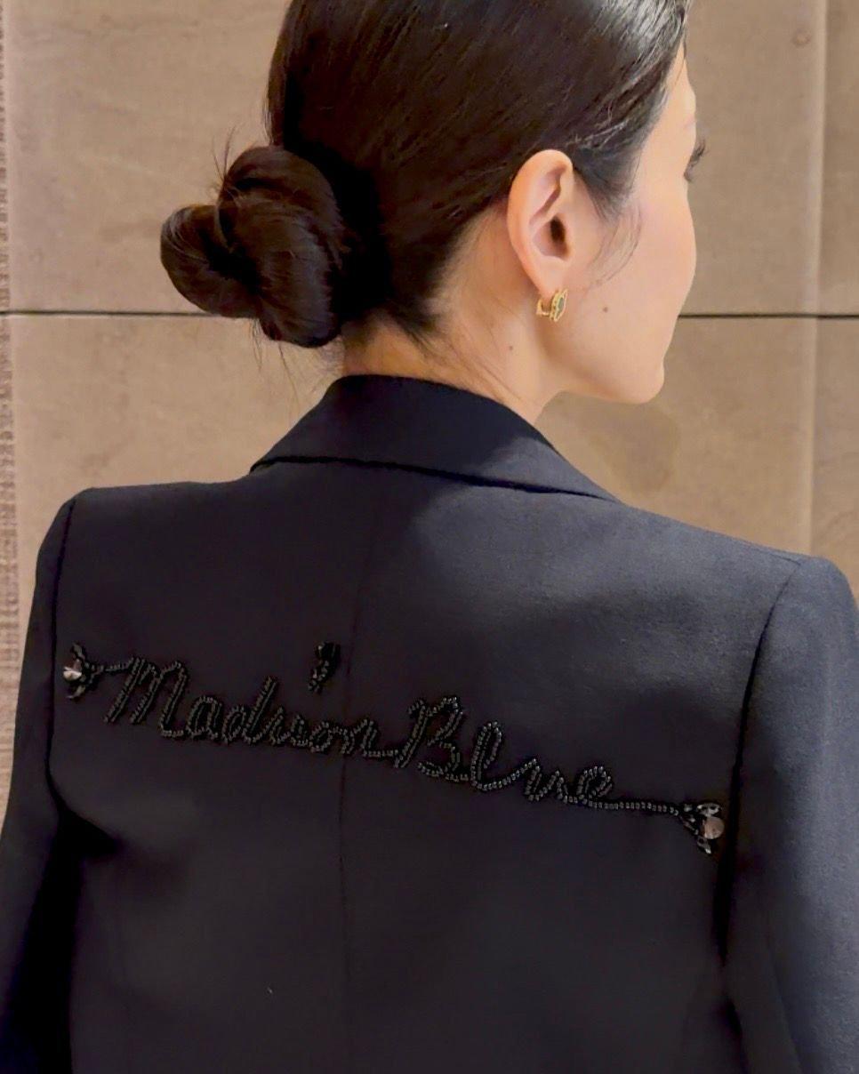 黒いジャケットの背面中央に、黒いビーズで「Madison Blue」と刺繍されている。背中は光沢があり、襟はきちんと形作られている。モデルはダークブラウンの髪をまとめ、耳には小さなピアスをしている。
