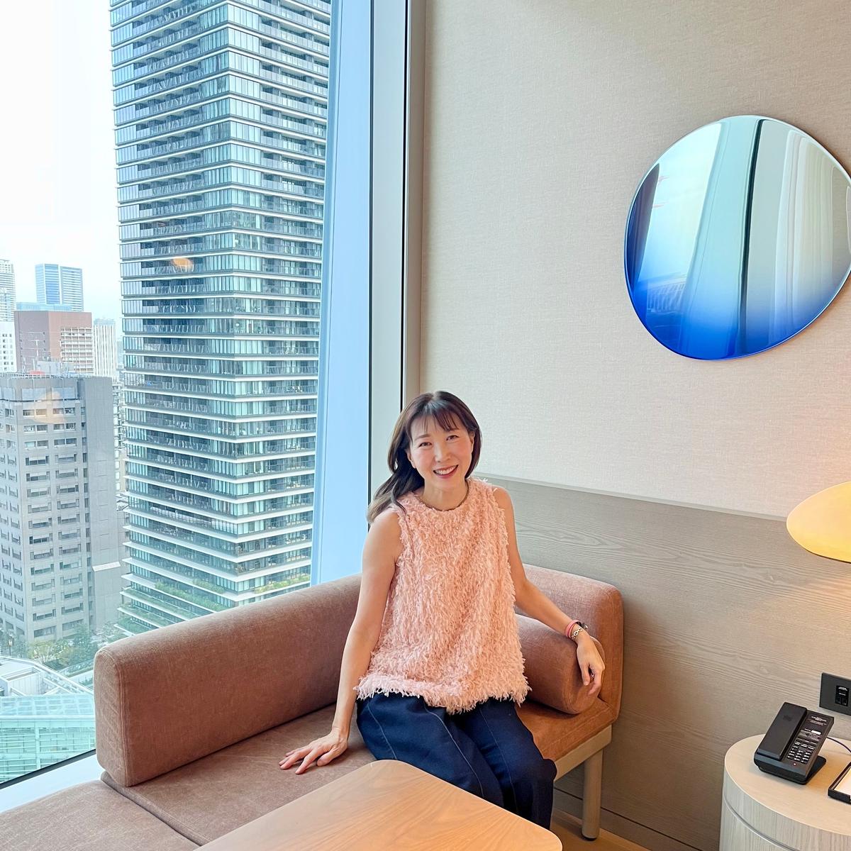 女性がベージュのふわふわしたベストと紺色のズボンで、窓のそばのベージュのソファに座っている。背景には高いガラスのビルが並び、丸い鏡と丸いテーブルが置かれている。