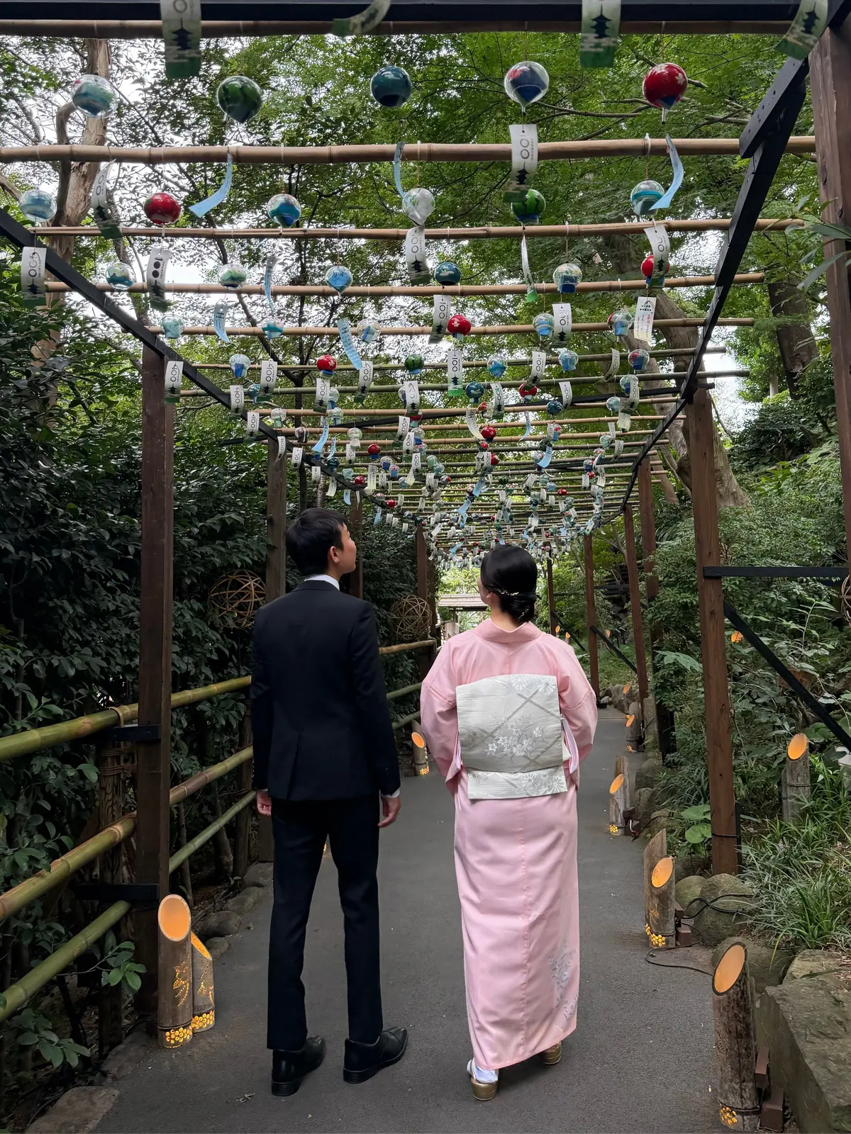 庭園の中を歩く新郎新婦。新郎は黒のスーツ姿、新婦はピンクの着物姿。頭上にはカラフルなガラス玉が多数吊るされ、幻想的な雰囲気を醸し出している。