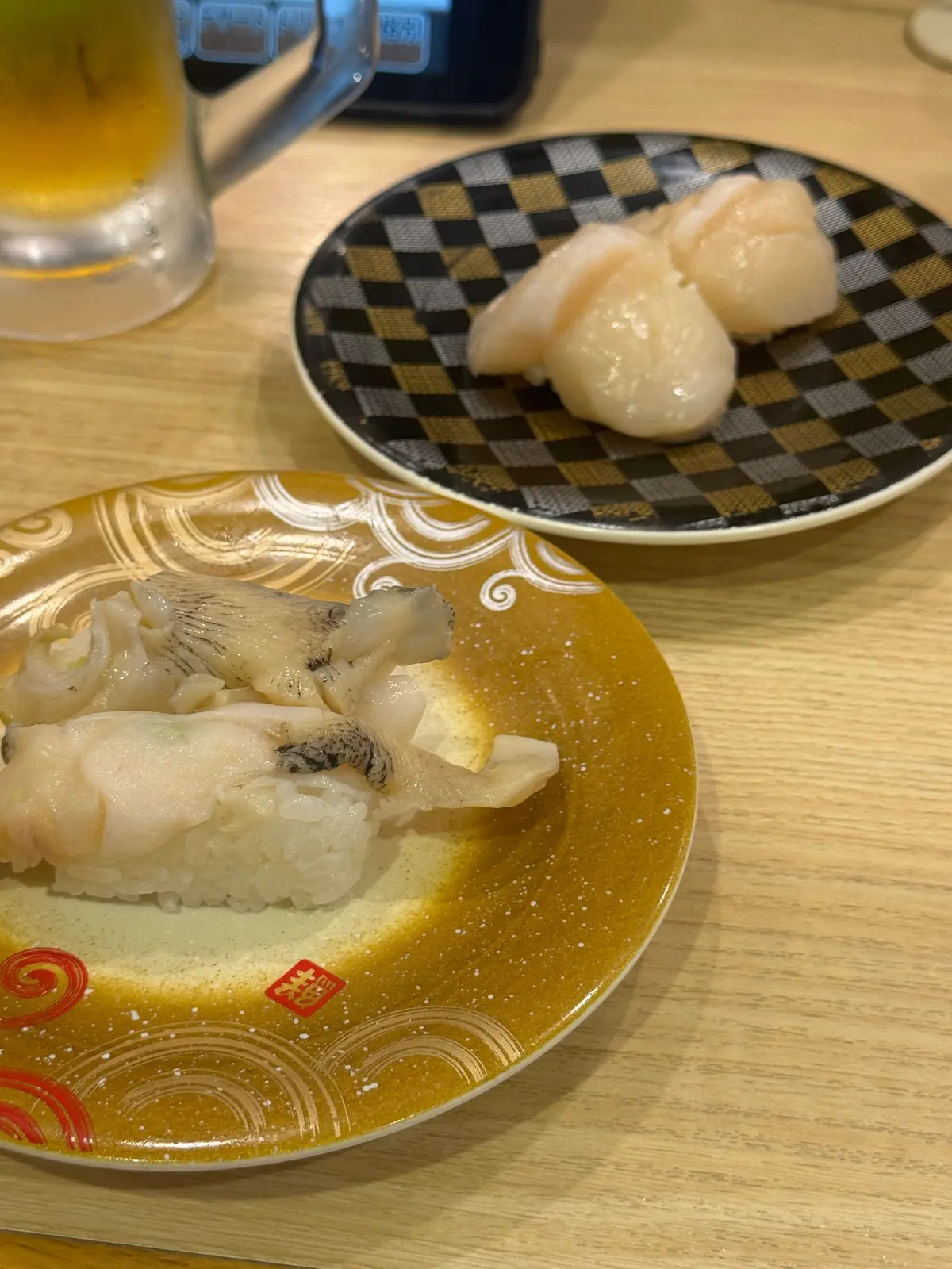 木製のテーブルに置かれた、寿司の盛り合わせ。手前は渦模様の皿にホタテの寿司が3切れ、奥にはチェック柄の皿にサーモンの寿司が3切れ。ガラスのグラスに入った飲み物と、スマートフォンが写っている。