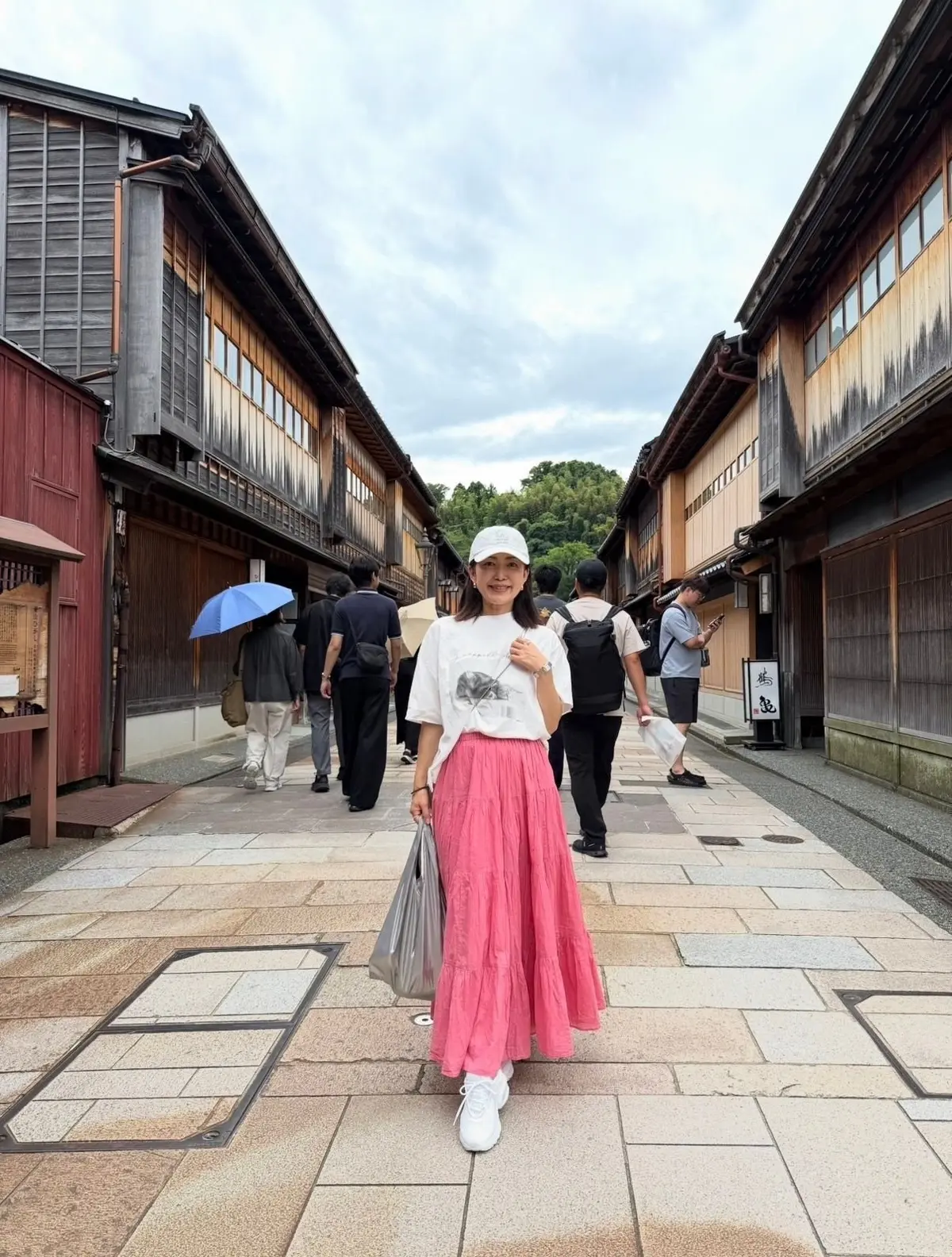 白帽を被った女性が、ピンクのスカートと白のTシャツで石畳の道に立っている。背景には木造の建物が並び、数人の歩行者がいる。