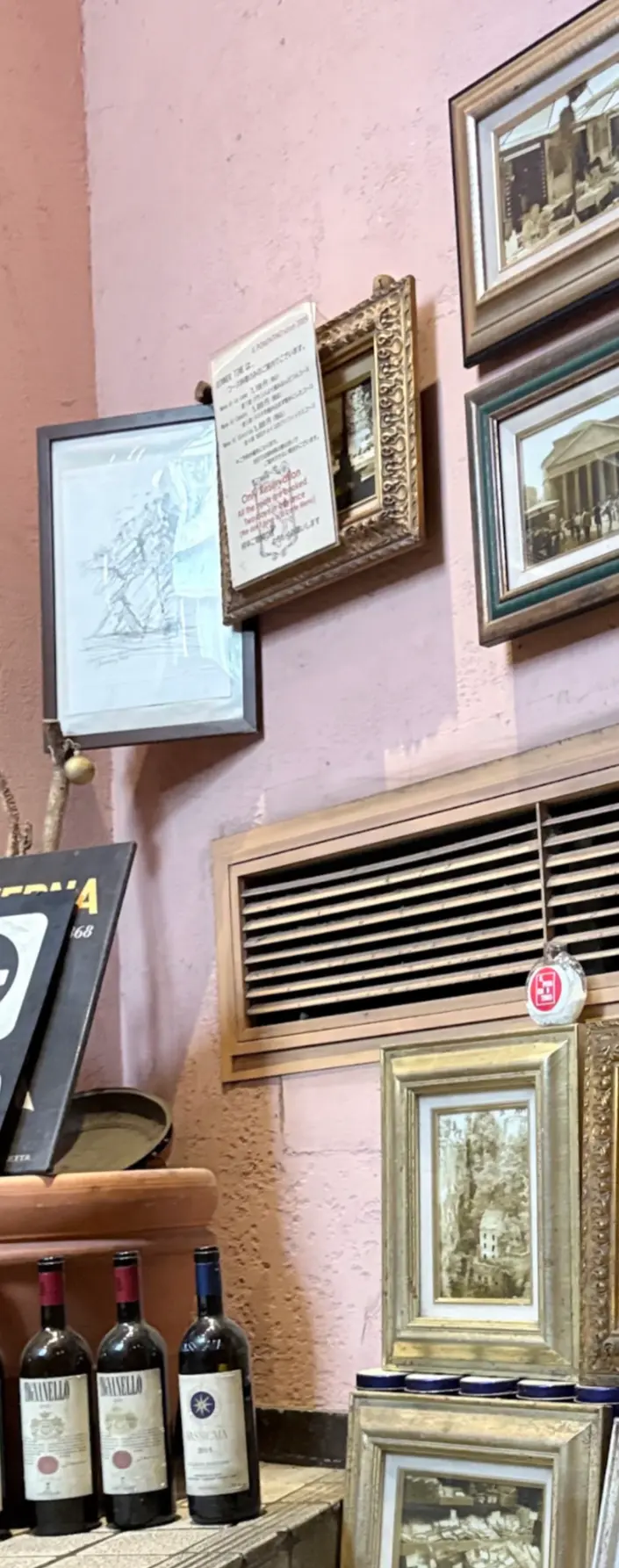 ピンクの壁にさまざまな額装された絵画が並び、棚には複数のワインボトルと青いグラスが置かれている。