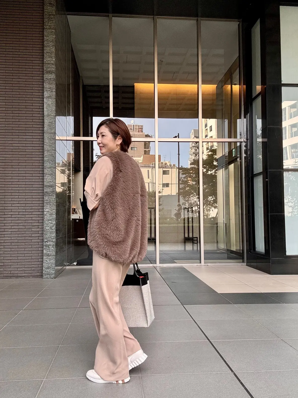 ベージュのファーベストとパンツルックを着た女性が、ガラス張りの建物の前を歩いている。右手にショッピングバッグを持ち、後ろ姿をこちらに向けている。背景には、住宅街の風景が映り込んでいる。