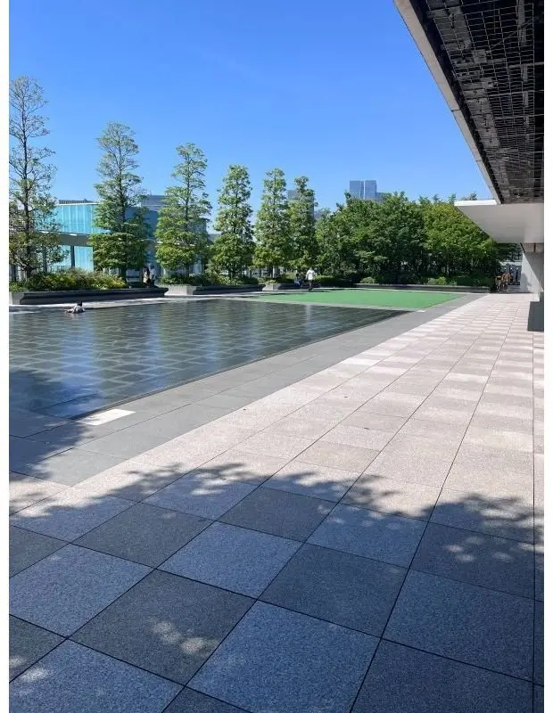 モダンなコンクリート敷きの広場に、幾何学模様の水面が広がり、背景には緑豊かな木々や建物が並ぶ