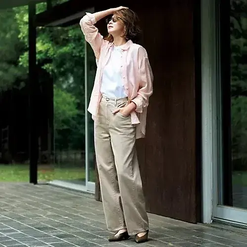 ベージュのパンツとピンクのシャツを合わせた女性。右手を頭に当て、サングラスをかけている。背景には木々が生い茂る緑豊かな風景と、モダンな建物の木製フレームが見える。