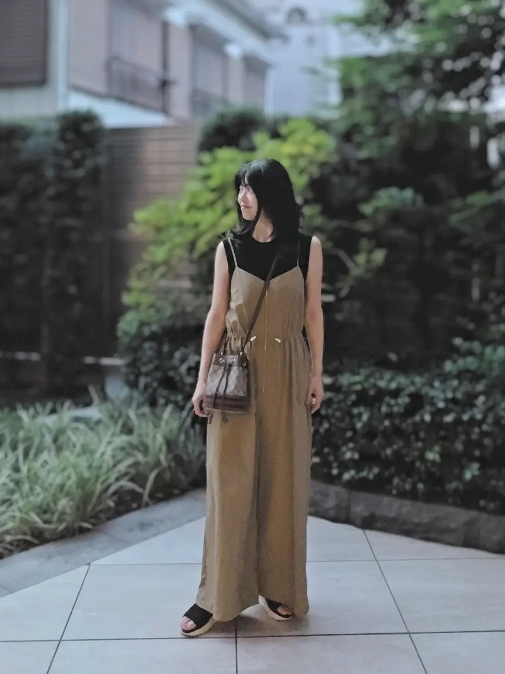 黒いTシャツの上にベージュのキャミソールワンピースを着た女性。右手に茶色のバッグを持ち、黒いサンダルを履いている。背景には緑の植物と白い建物が見える。