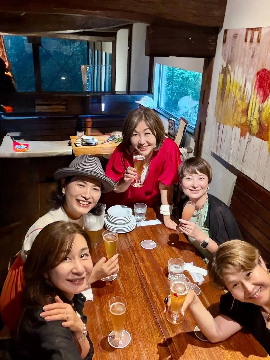 木製のテーブルを囲み、笑顔の女性6人がビールグラスを持ちながら談笑している様子。背景には窓から緑が見える。
