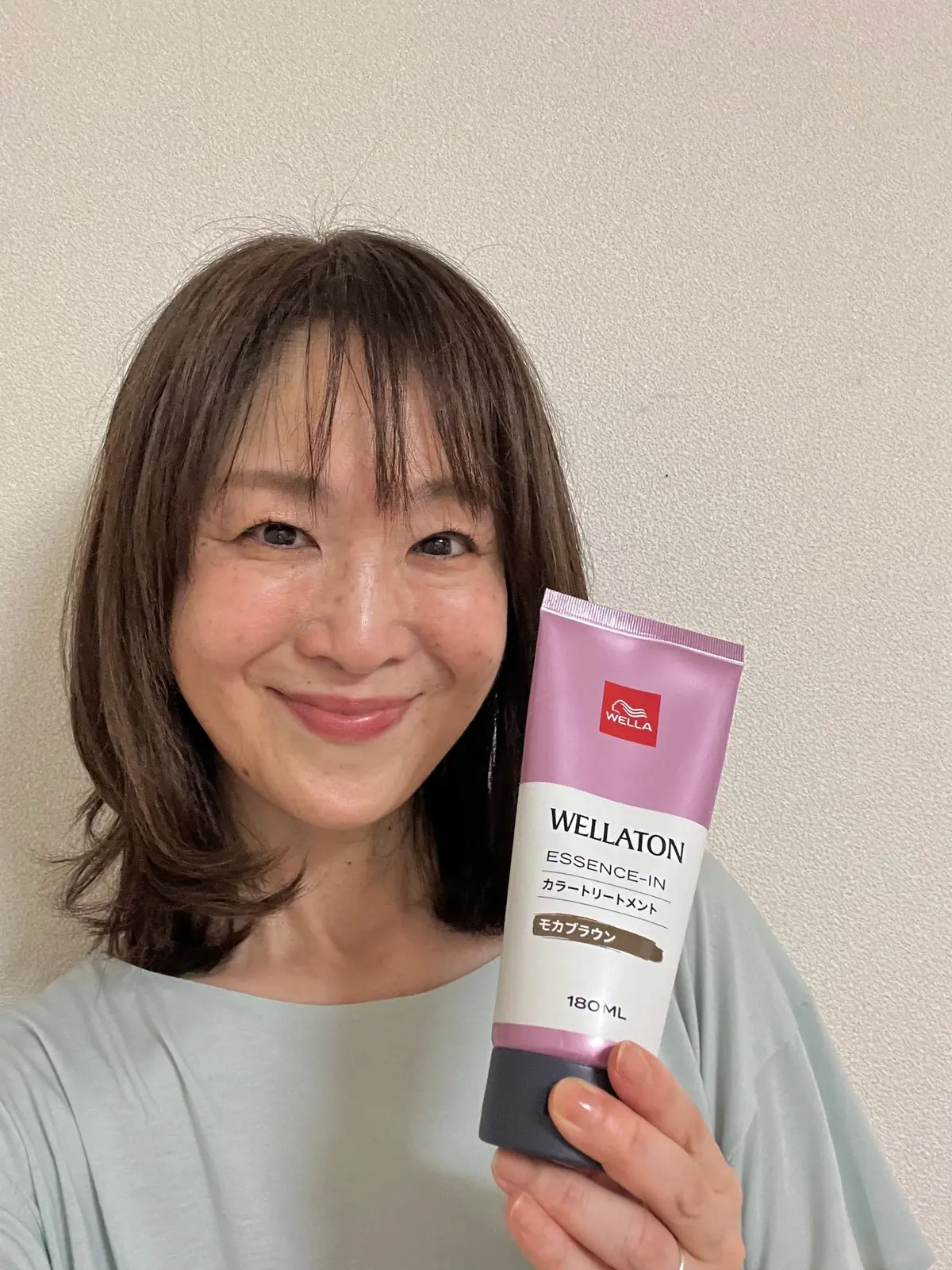 笑顔の女性が、白い壁の前でウェラトーンのエッセンスインカラートリートメントのチューブを持つ。茶色のショートヘアで、白いTシャツを着ている。