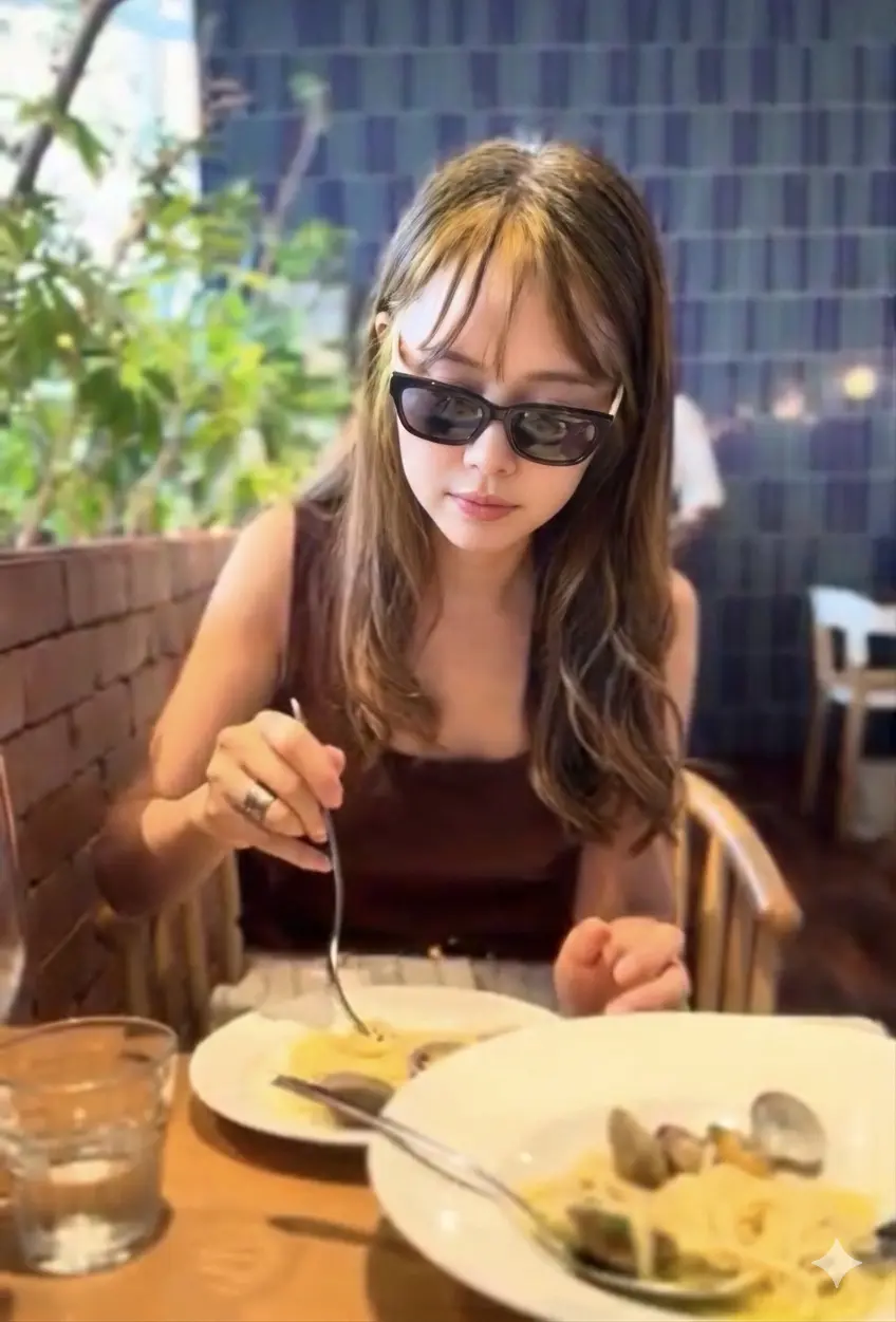 女性がサングラスをかけ、木製のテーブルに置かれたパスタと貝類の料理をフォークで食べている。背景には緑の植物と青いタイルの壁が見える。
