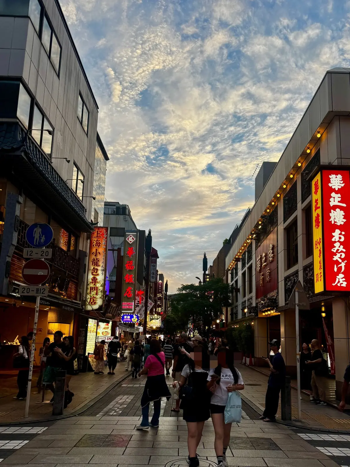 夕暮れ時の商店街の風景。両側に建物が並び、ネオンサインが輝いている。多くの人が歩き、活気がある。空は雲が多く、オレンジ色と青色のグラデーション。