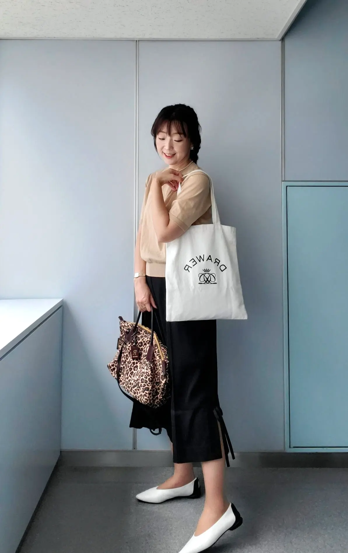 ベージュのキャンバストートとレオパード柄のバッグを持つ女性。グレーのニットの上に淡いピンクのシャツを着用し、黒いタイトスカートと白いローファーを履いている。背景には白と水色の壁がある。