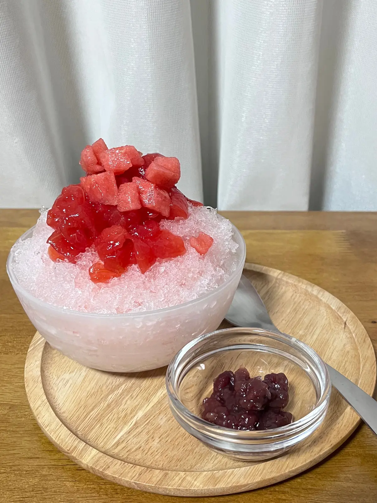 木製のトレーに置かれた、ピンク色の氷の上にカットされたスイカが盛り付けられたデザート。小さなガラス器に入った紫色の豆菓子が添えられている。背景には白いカーテン。