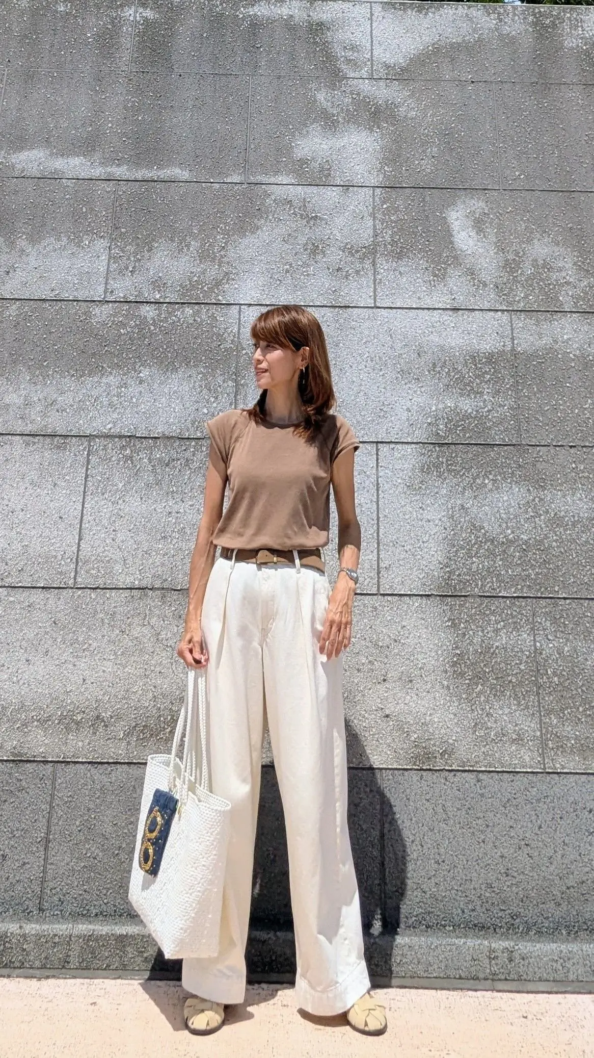 茶色のカットソーに白いワイドパンツを合わせた女性。白いトートバッグを提げ、グレーの壁の前でポーズをとっている。