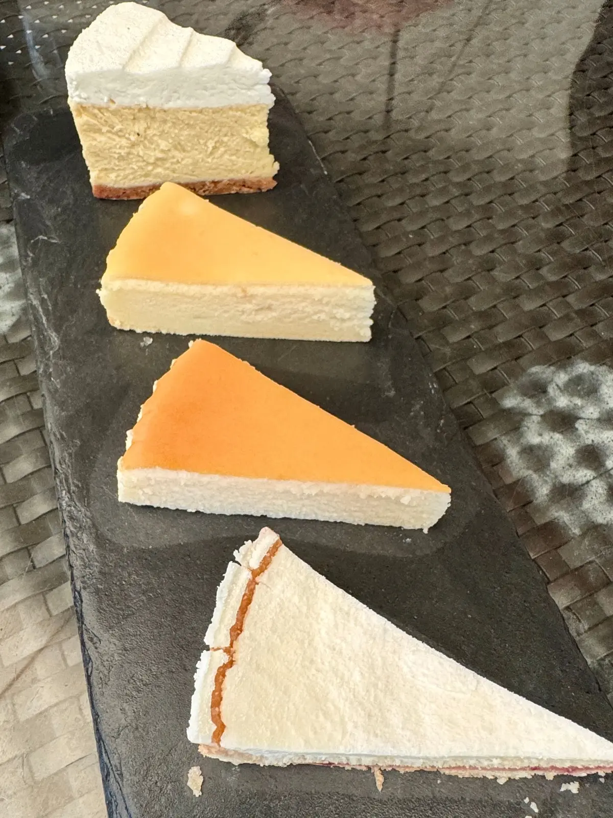 黒い石板の上に、4切れのチーズケーキが並んでいる。上から、白いクリームチーズ、オレンジ色のマンゴー、オレンジ色のパッションフルーツ、プレーンのチーズケーキ。