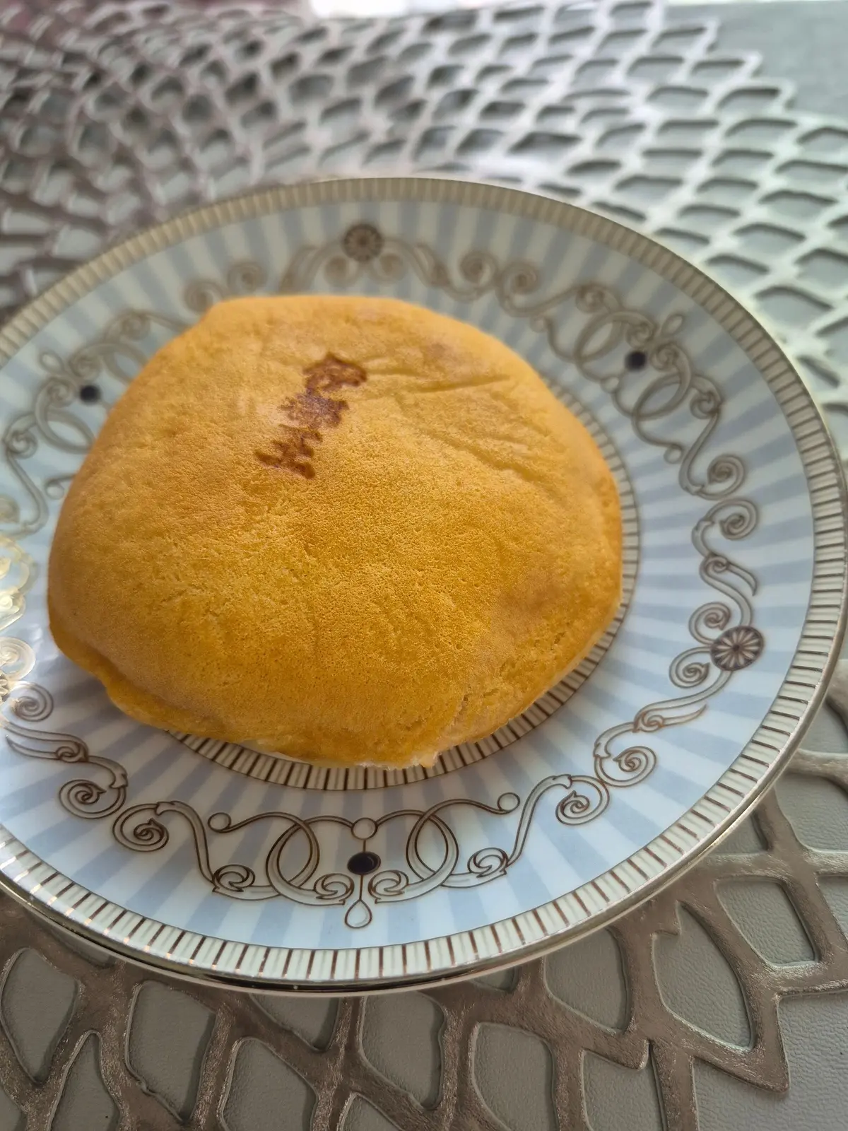 黄色の丸いパンが、淡い青色の模様の皿に置かれている。パンの上には、茶色の文字が刻まれている。背景には、白いレースのような模様が見える。