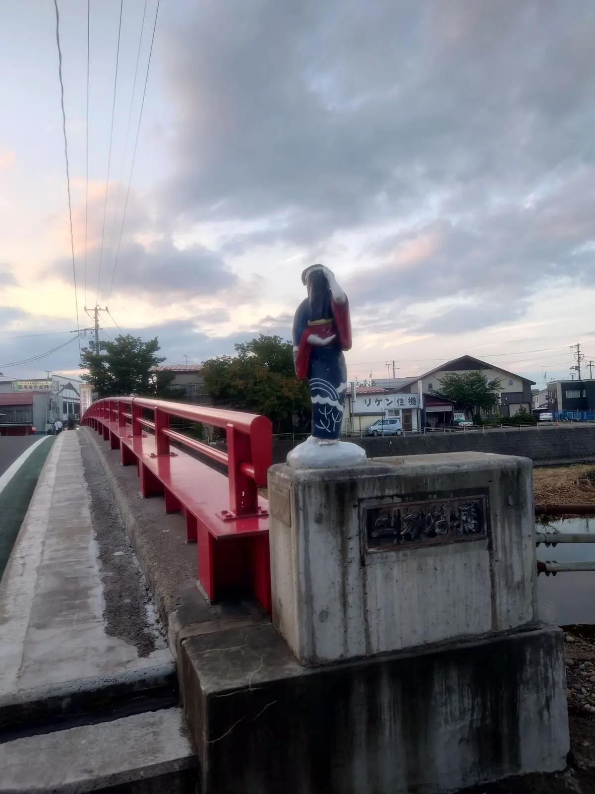 赤い橋の欄干に置かれた、装飾的な鳥の像。背景には住宅街と夕焼け空が広がり、穏やかな雰囲気。