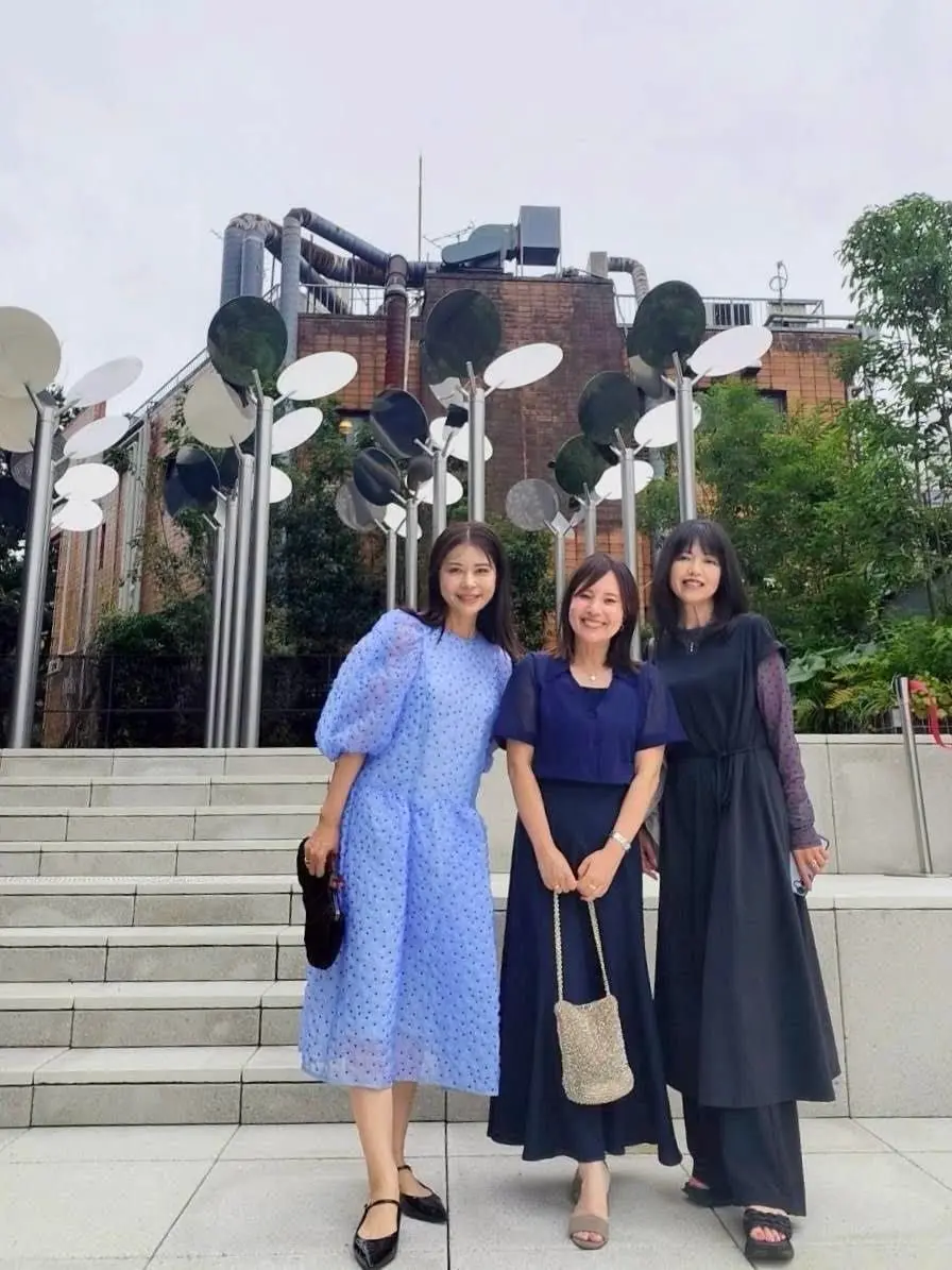 3人の女性が階段の前に並んで立っている。左から青色のドット柄ワンピース、紺色のブラウスとスカート、黒いワンピースを着ている。背景には、金属製のパイプから円盤状のオブジェが吊り下げられた現代アート作品と、レンガ色の建物がある。