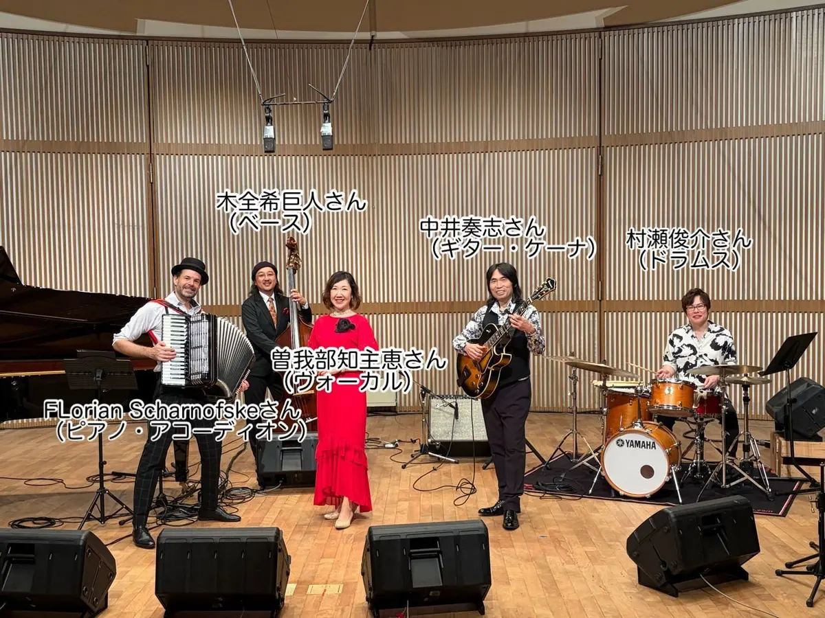 ステージ上で楽器を演奏する5人のミュージシャン。左からアコーディオン、ベース、ヴァイオリン、ギター、ドラム。背景にはグランドピアノと観客席が見える。